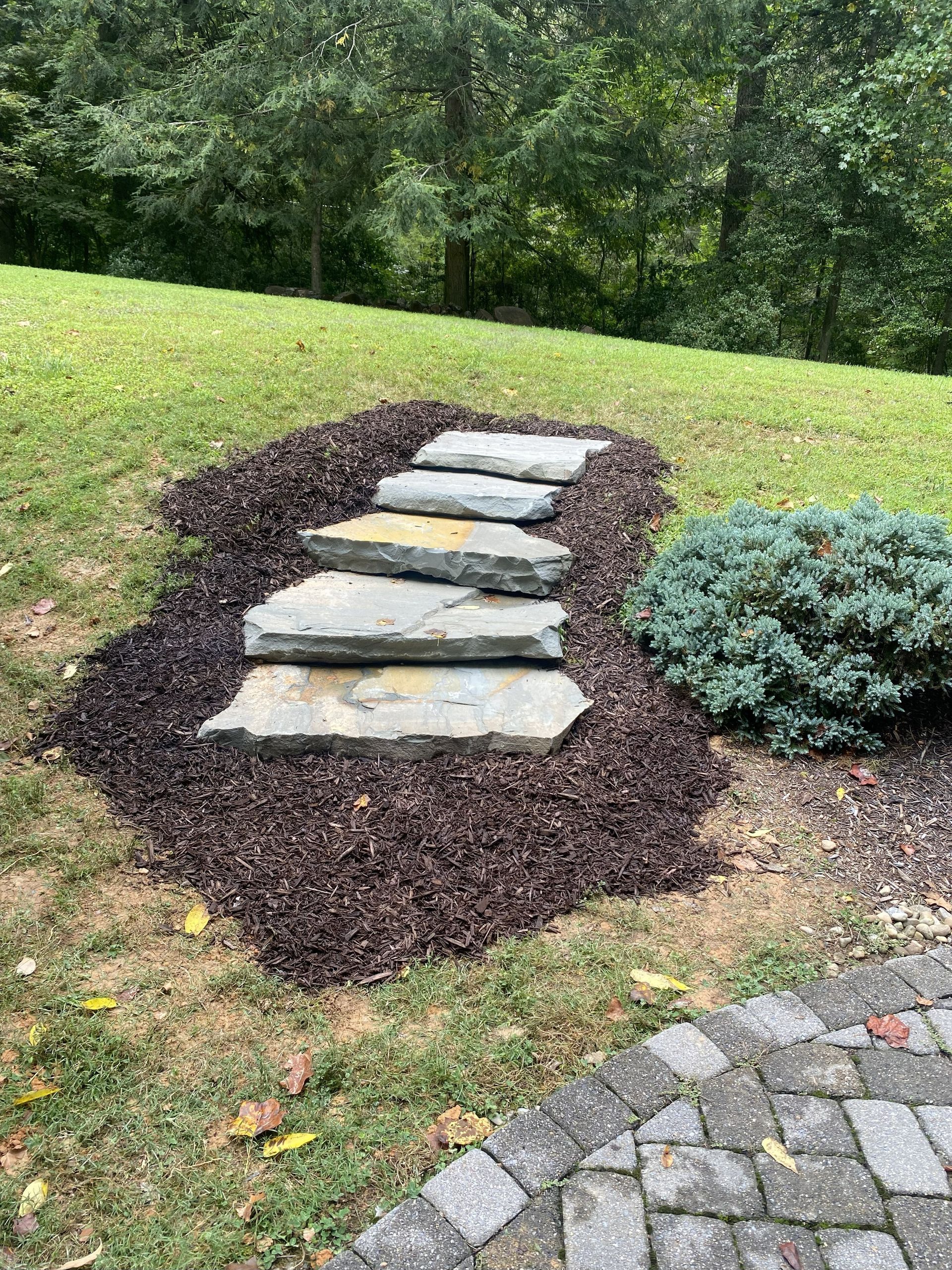 Stone steps set in a bed of dark mulch on a grassy lawn near a paved stone walkway.