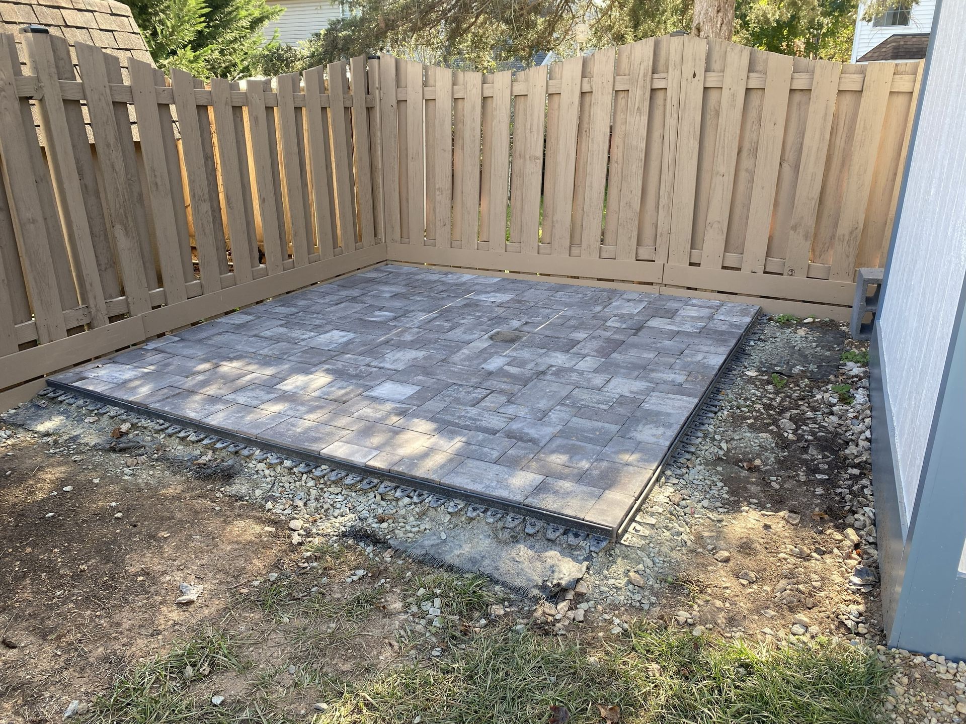 A new square patio made of grey rectangular pavers, situated in a corner yard with a tan wooden fence.