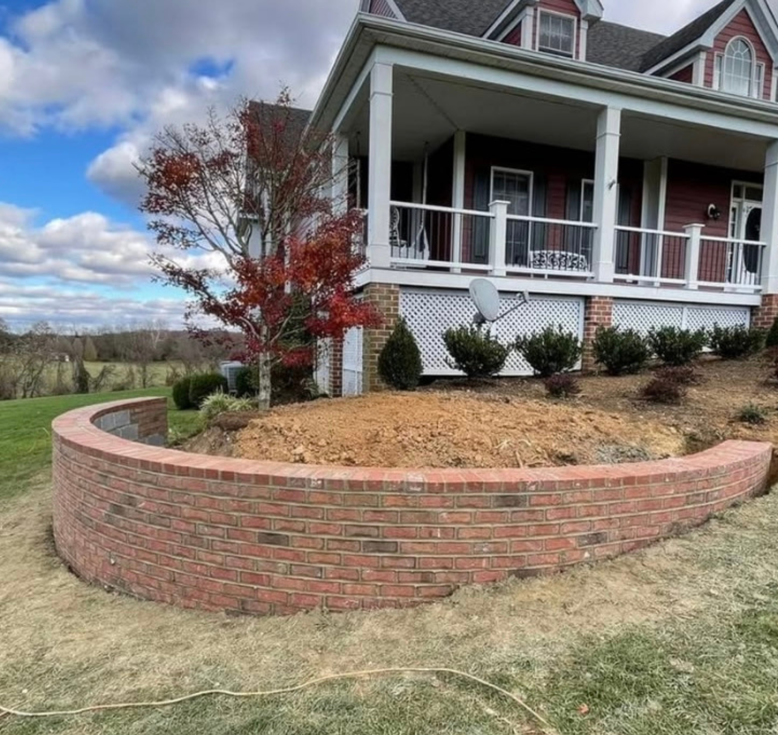 A curved red brick retaining wall in a yard in front of a red farmhouse with a porch under a blue sky with clouds.