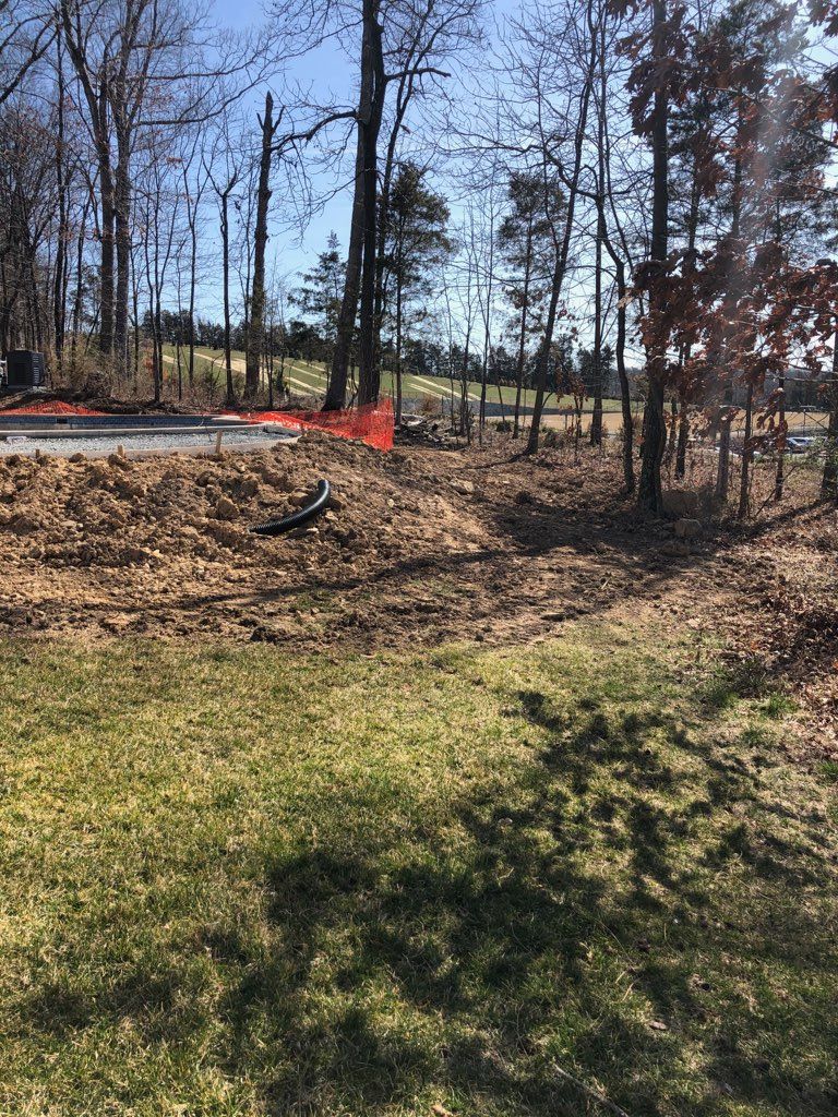 A construction site with a dirt mound, orange safety fencing, and a black pipe in a wooded area near a grassy lawn.