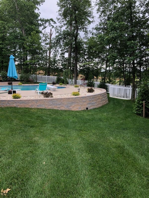An elevated backyard pool area with stone retaining walls, a blue umbrella, and a white fence surrounded by trees.