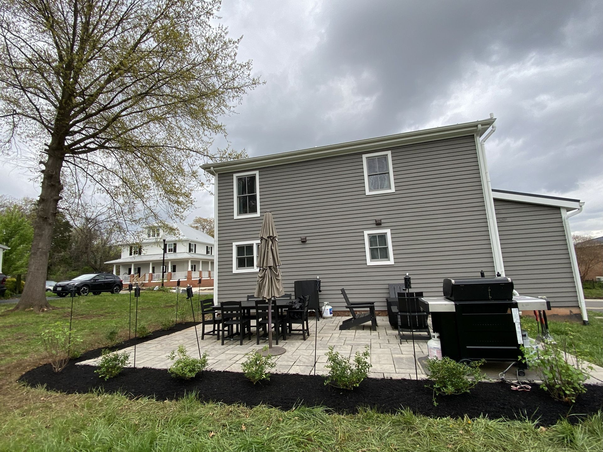 A grey-sided house with a stone patio featuring patio furniture and a grill, set against a lawn and cloudy sky.