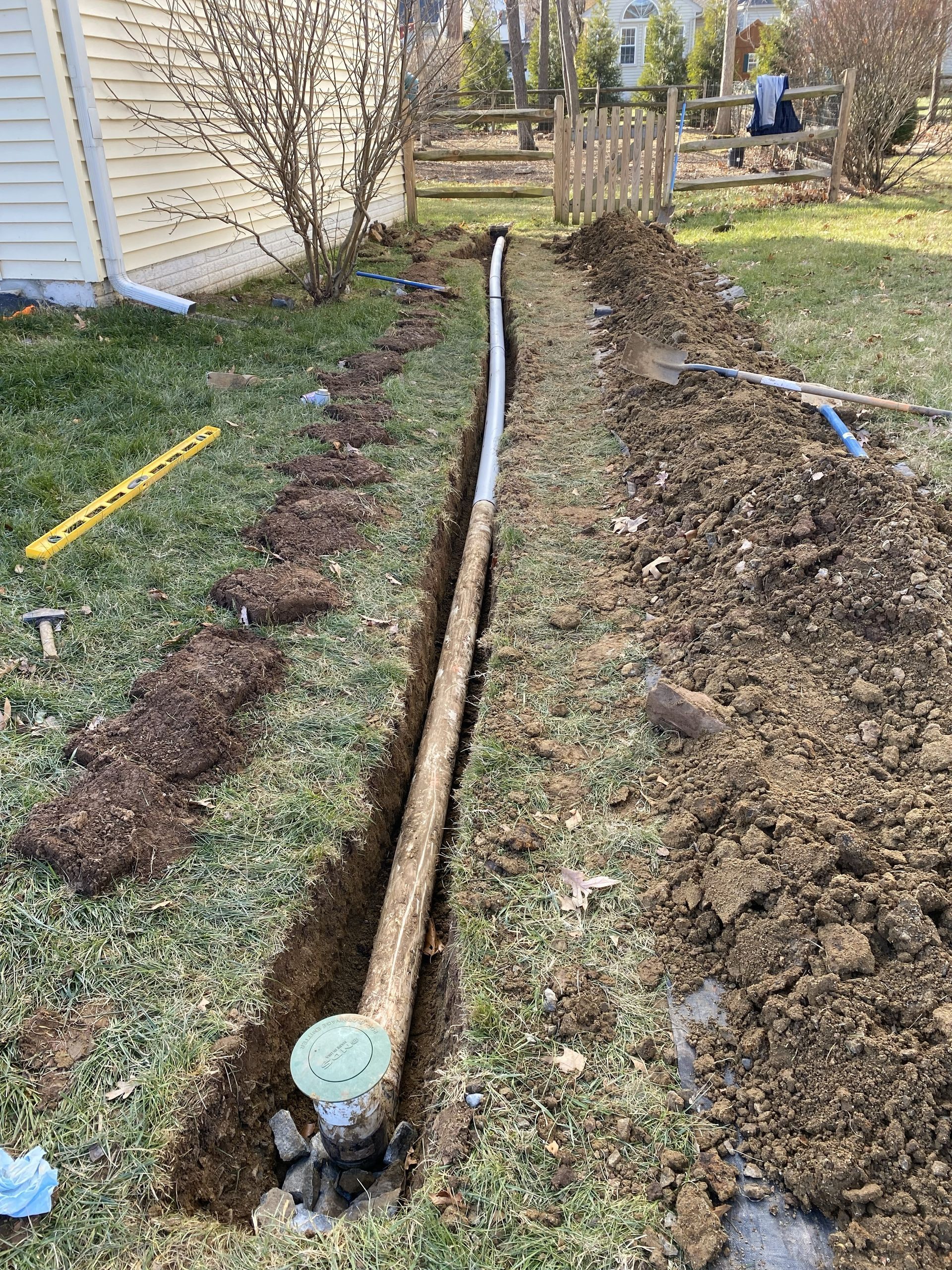 A trench in a grassy yard showing a newly installed sprinkler pipe and valve box along the side of a house.