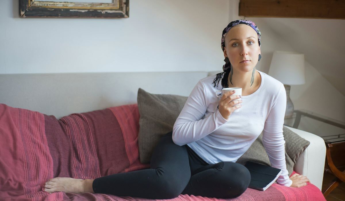 Woman sitting on a couch, holding a cup, with a pink blanket in a bright living room.