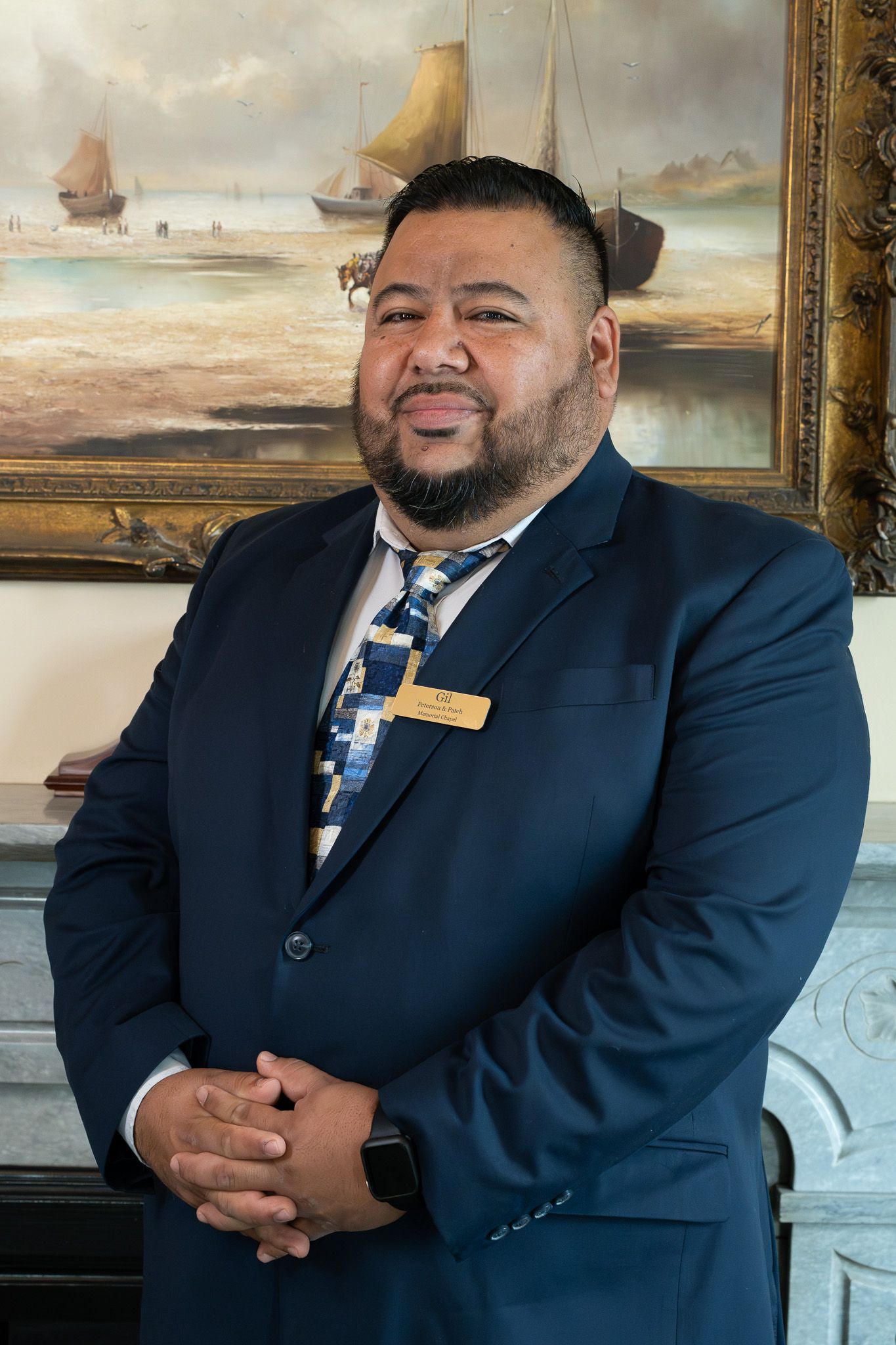 Man in a navy suit with gold tie clip, hands clasped, standing before a framed seascape painting
