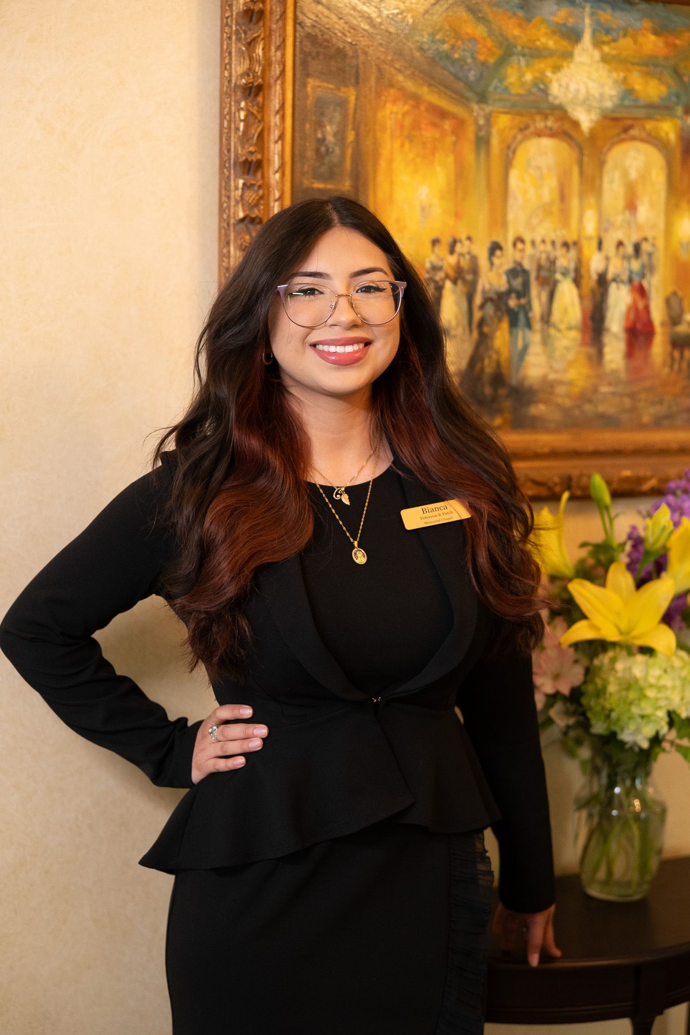 Woman in black standing indoors beside flowers and a framed painting, smiling at the camera.