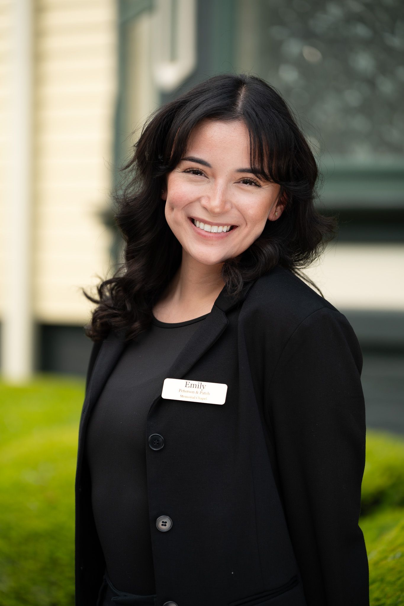 Smiling woman in a black blazer standing outdoors near a building.