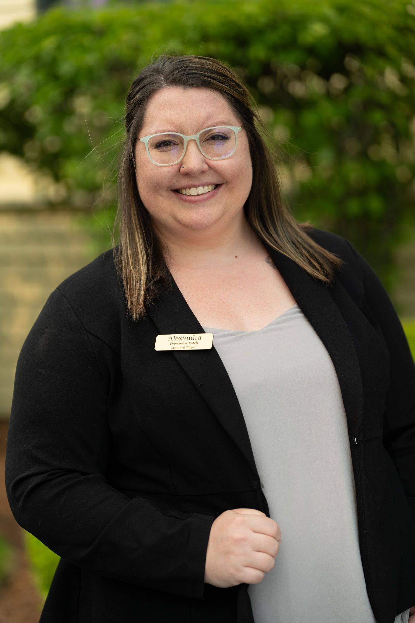 Smiling woman in glasses wearing a black cardigan and gray top, standing outdoors in front of greenery