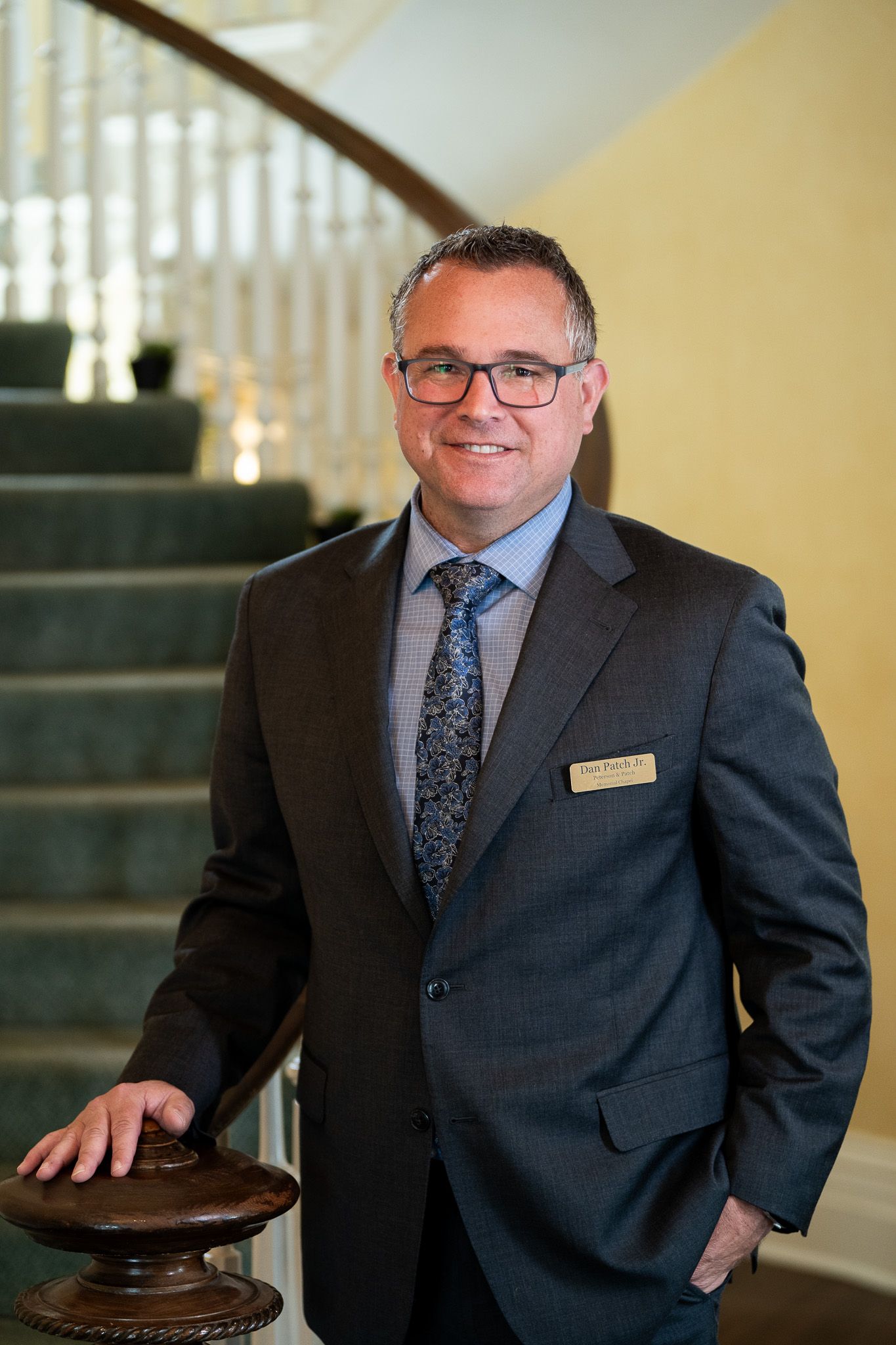 Man in a dark suit standing by a staircase, smiling with one hand on a wooden banister.