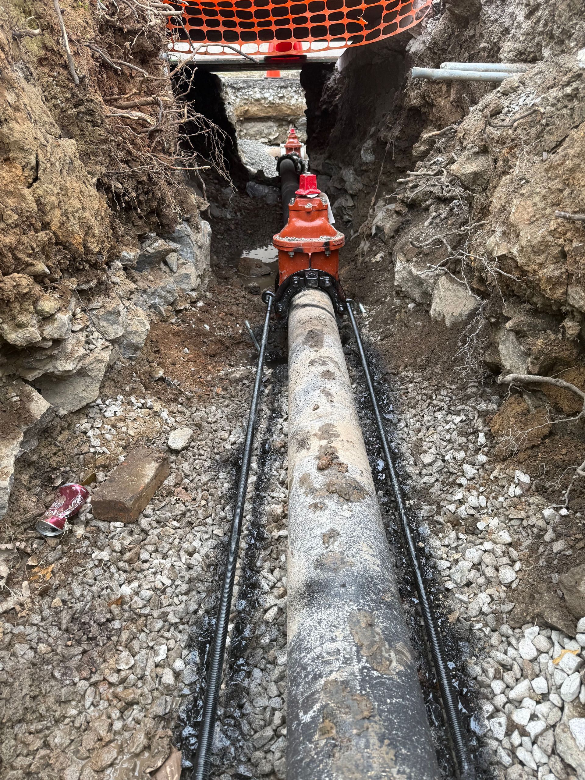 A bright orange fire hydrant is installed on a utility pipe in a narrow, excavated dirt trench.