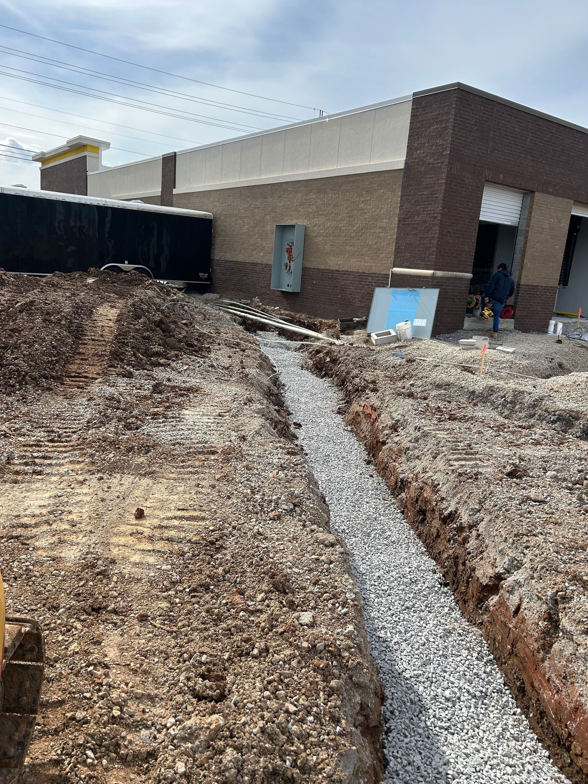 Construction trench beside a brick building, with gravel path and dry dirt in the foreground.