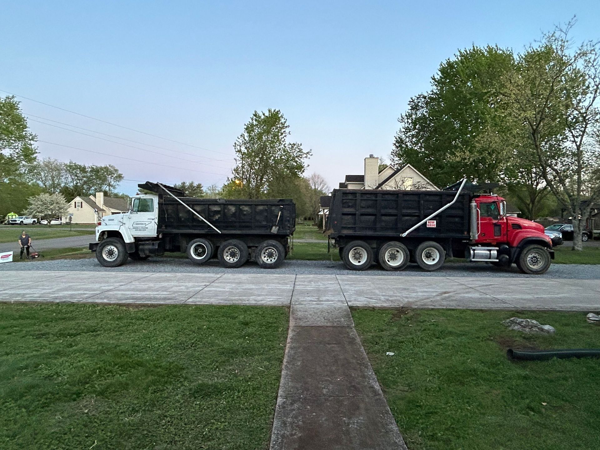 A white truck and a red truck with black dump trailers parked side-by-side on a gravel driveway in a grassy area.