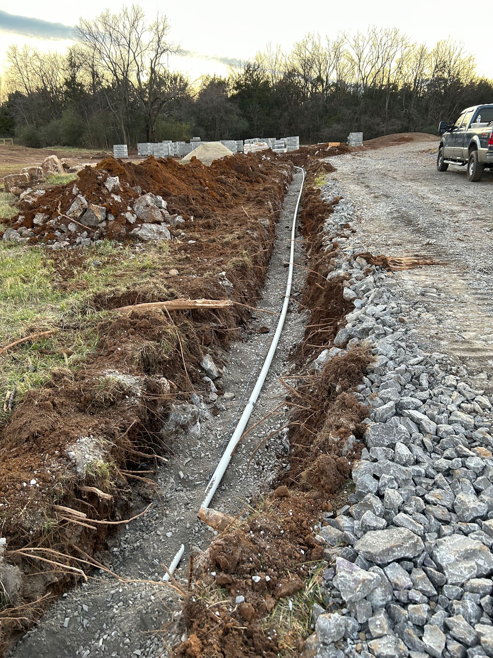 A long trench dug into dirt alongside a gravel road, with a pipe running down the center of the trench.