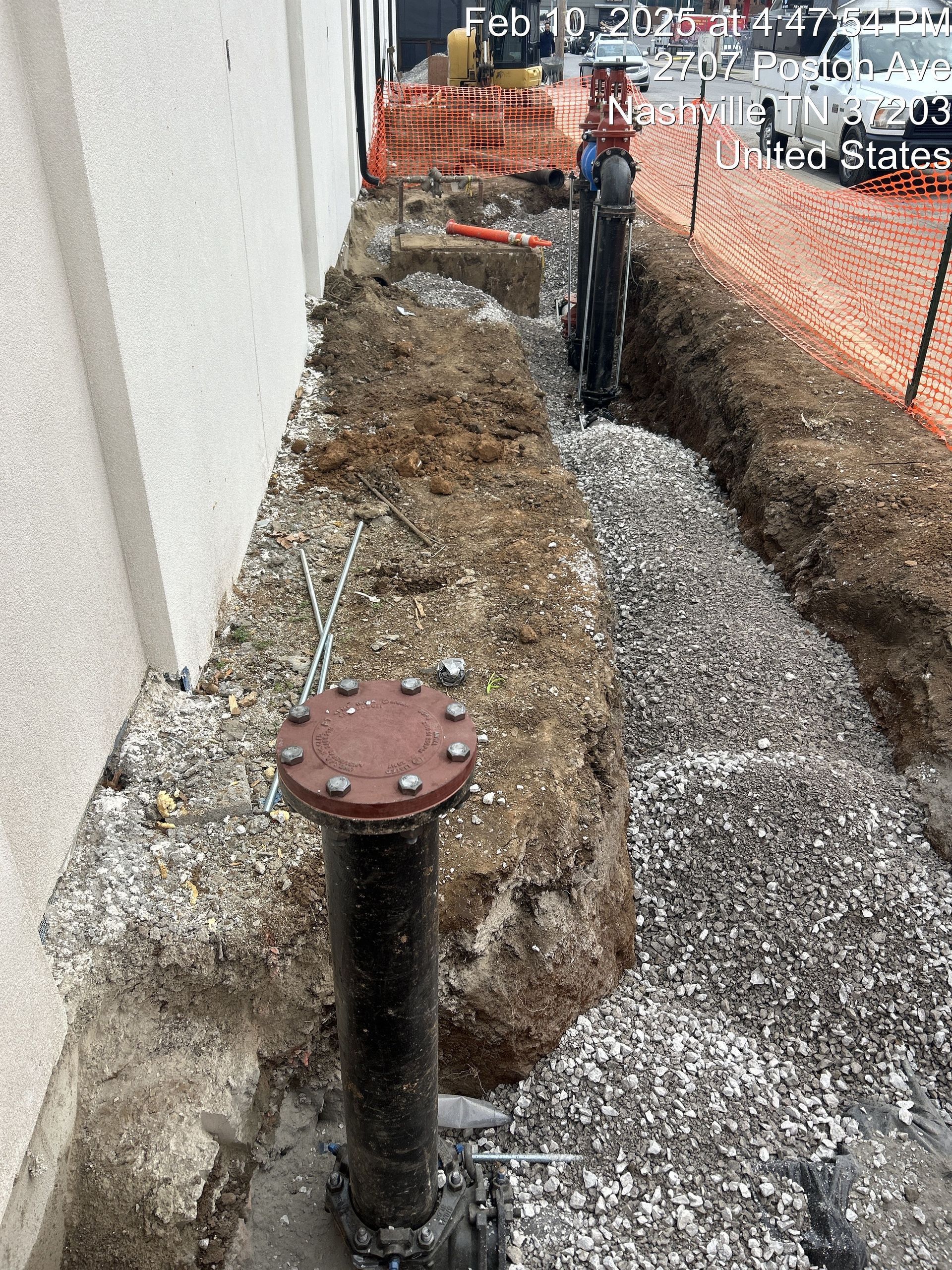 A black pipe with a bolted cap stands in a construction trench filled with gravel next to a building wall.
