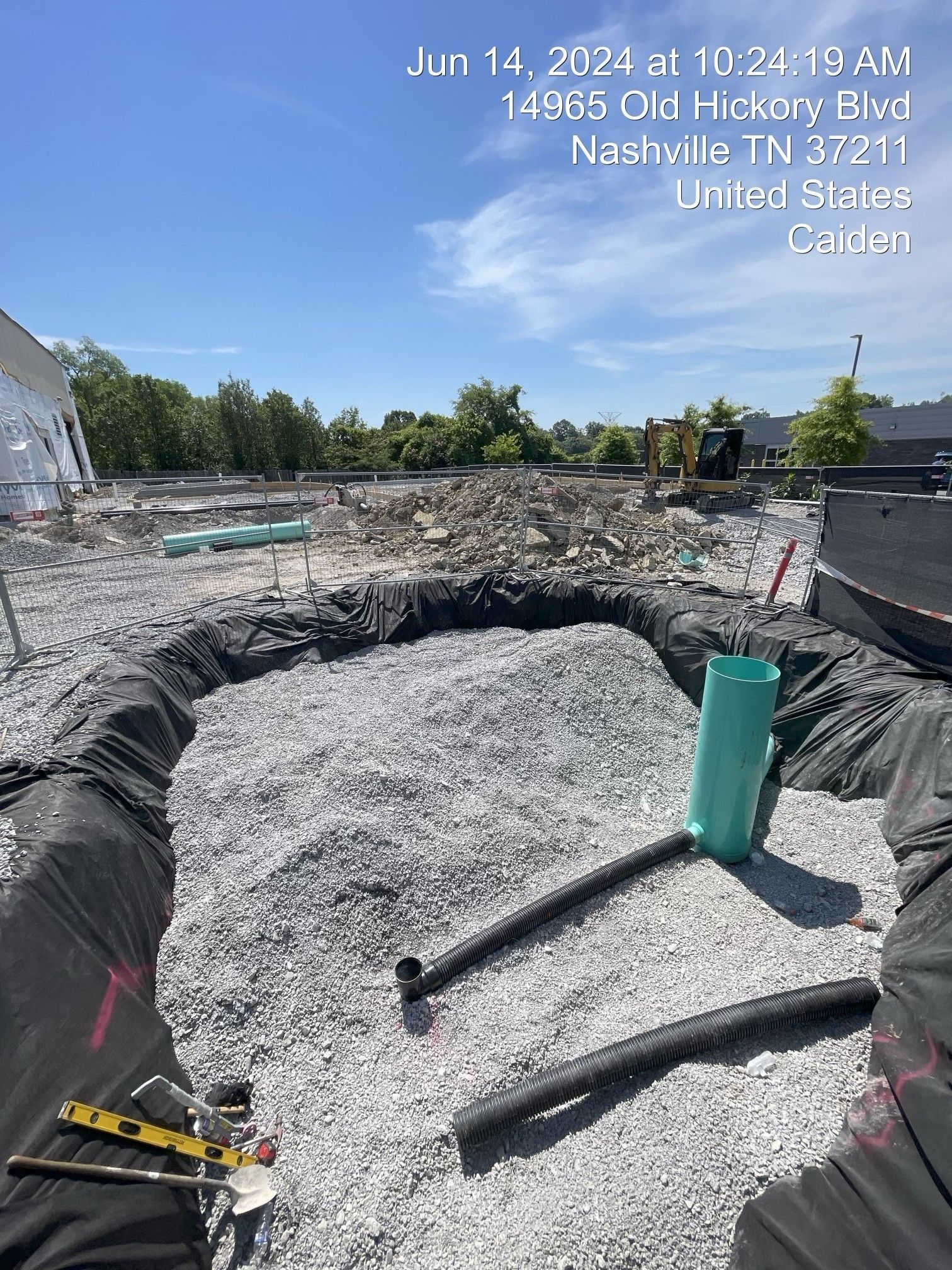 Construction site with a large excavated pit lined in black tarp and gray gravel, tools and pipes inside.