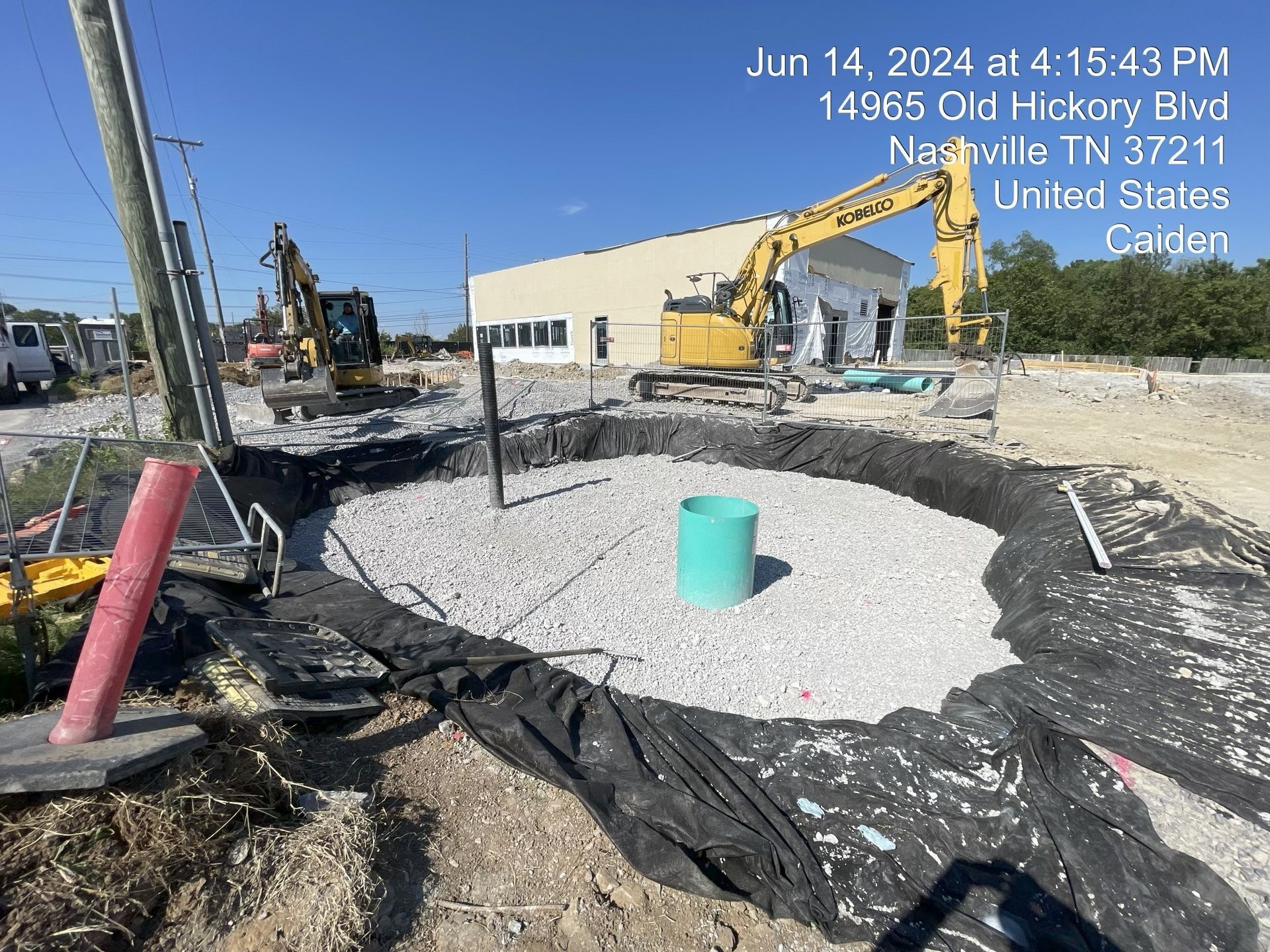 Construction site with excavator, black tarp-covered excavation, and a green bucket near a blue sky.