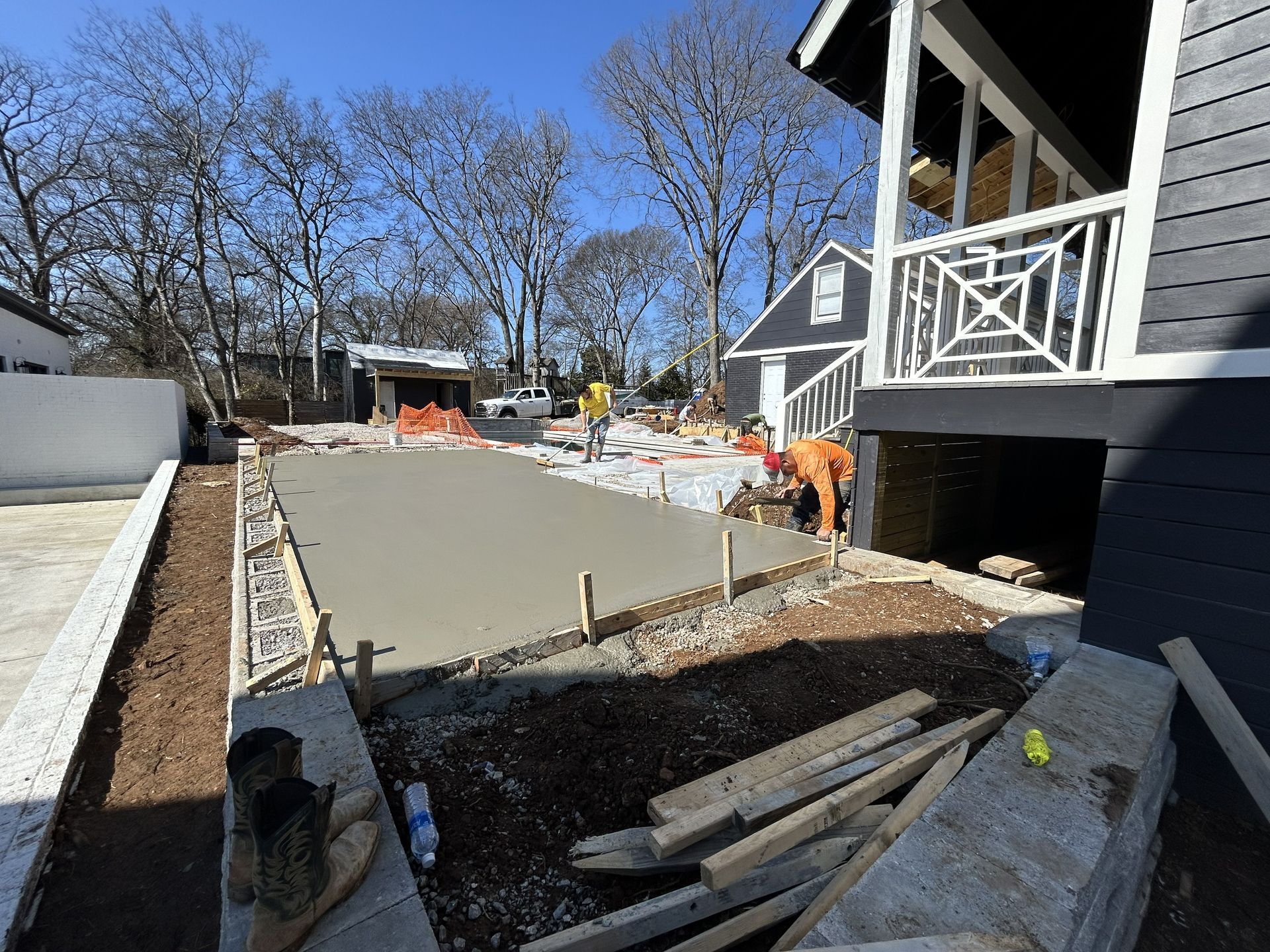 Concrete driveway under construction beside a gray house, with workers and tools in a sunny yard.