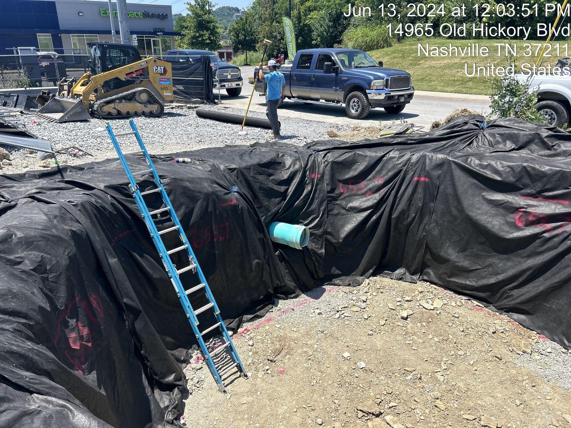 Excavation site with black tarp-lined pit, blue ladder, backhoe, and pickup trucks on a road.