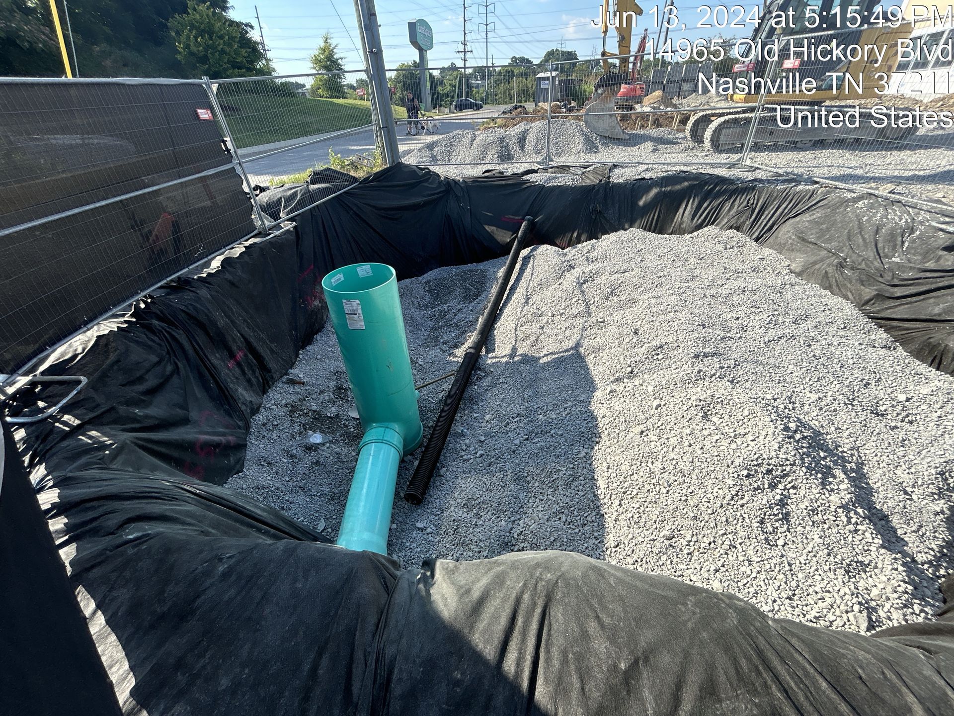 Open trench beside a road with black liner, gravel, and blue pipes under construction.