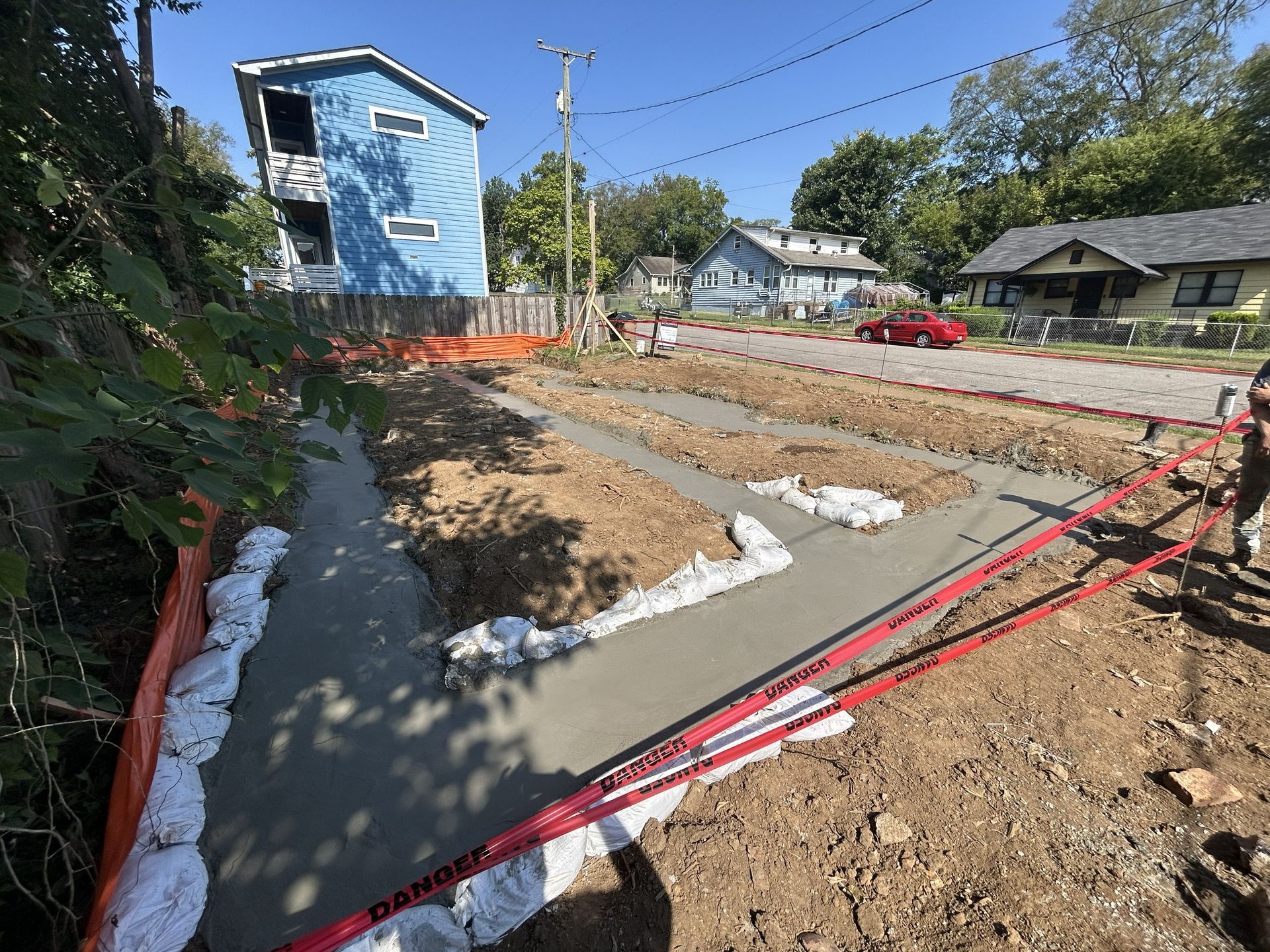 New sidewalk under construction beside a blue house, with red caution tape and sand piles.