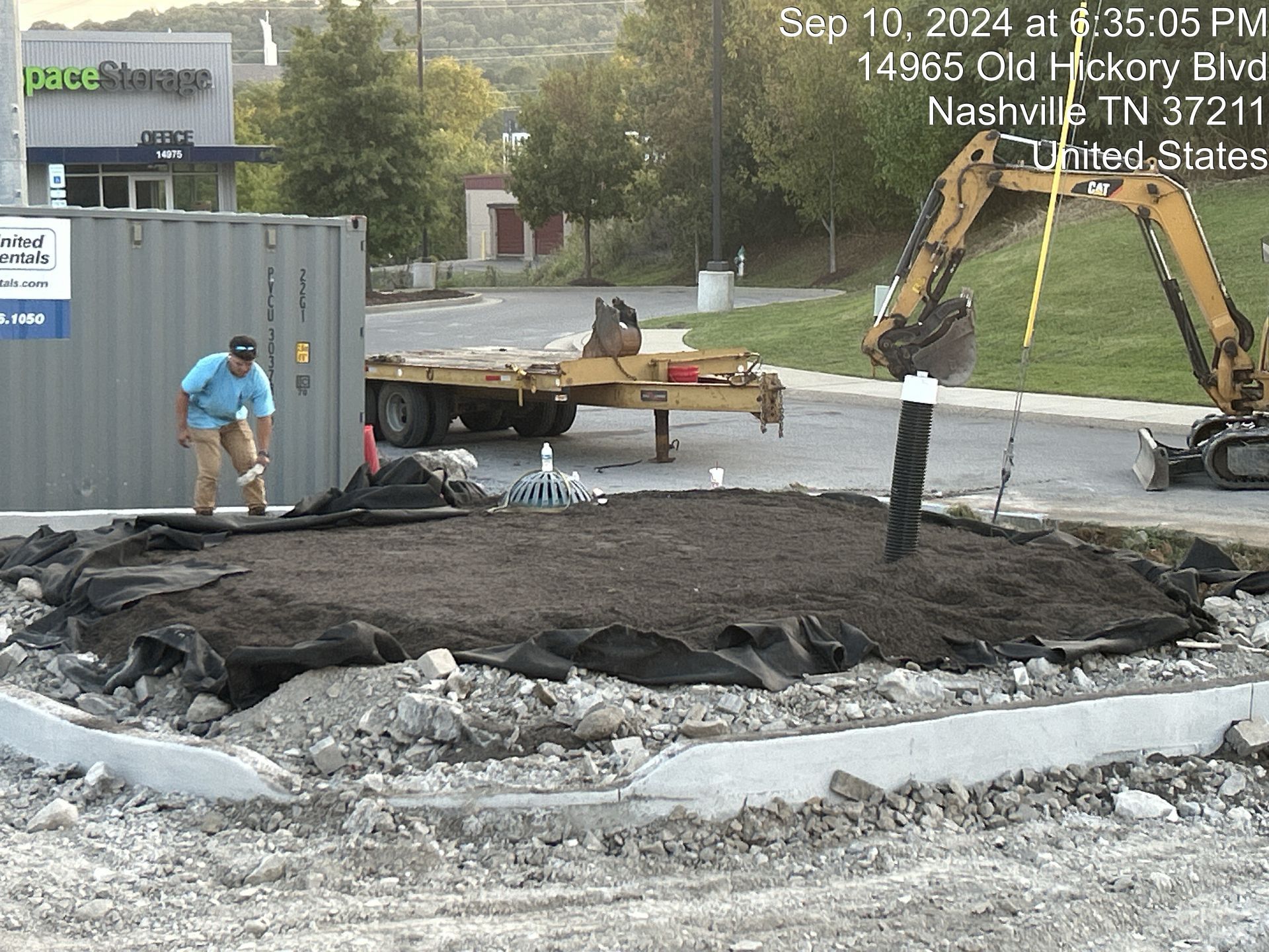 Construction crew repairing a torn-up road beside a gray container and excavator on a wet street