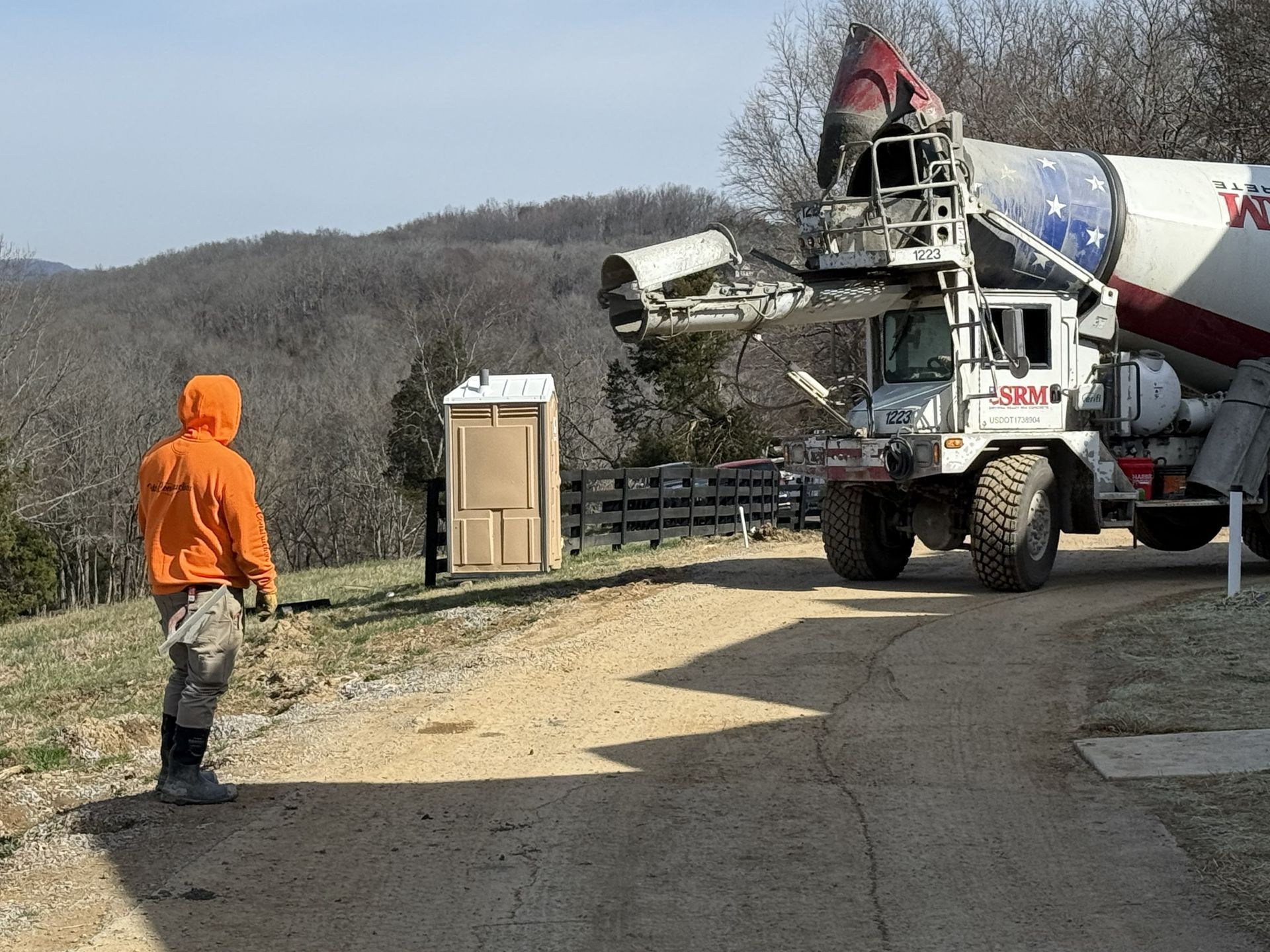 Cement mixer truck on a gravel road with a worker in an orange hoodie nearby, wooded hills in the background