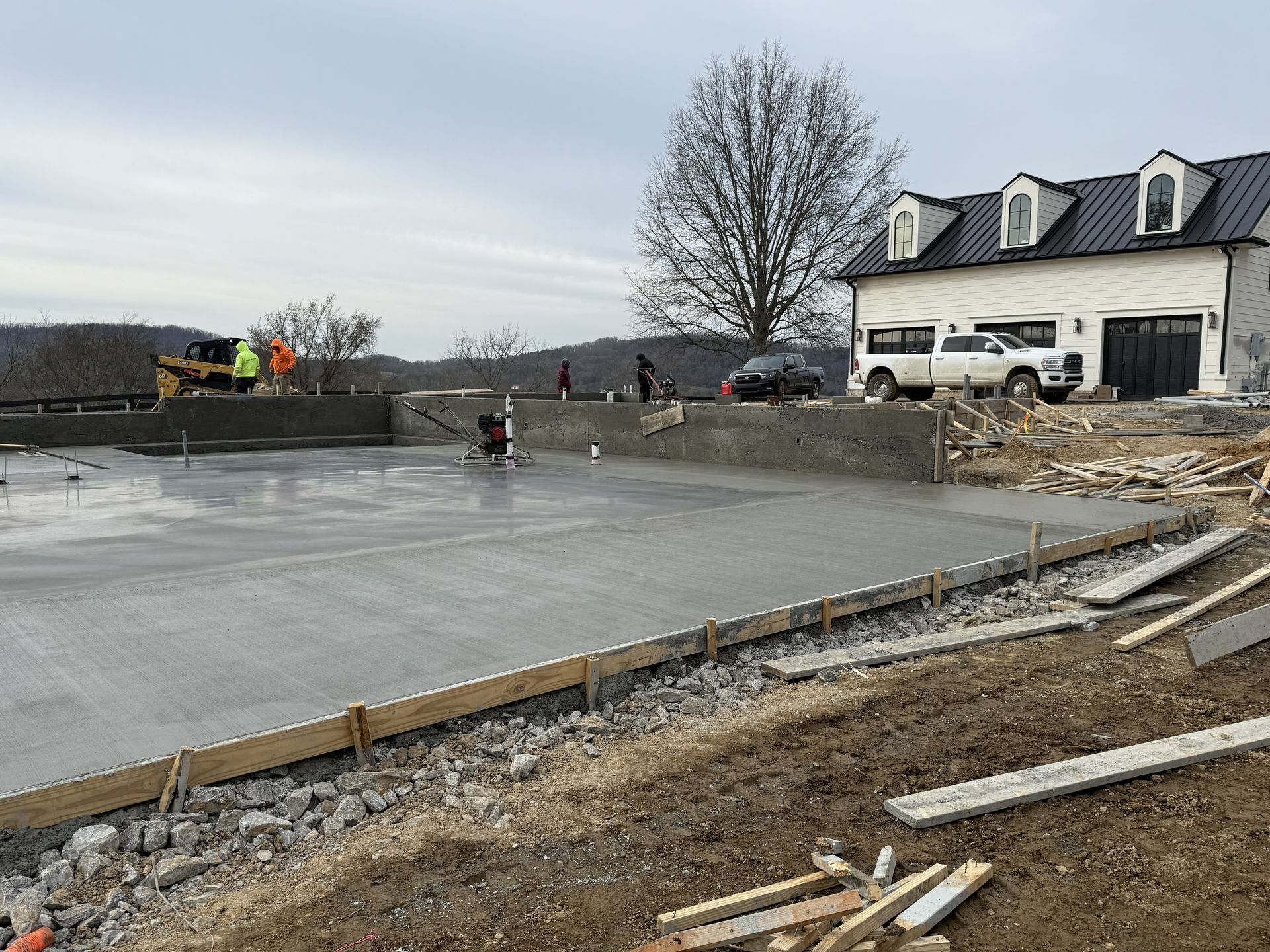 Fresh concrete slab being finished at a construction site beside a white house and leafless tree