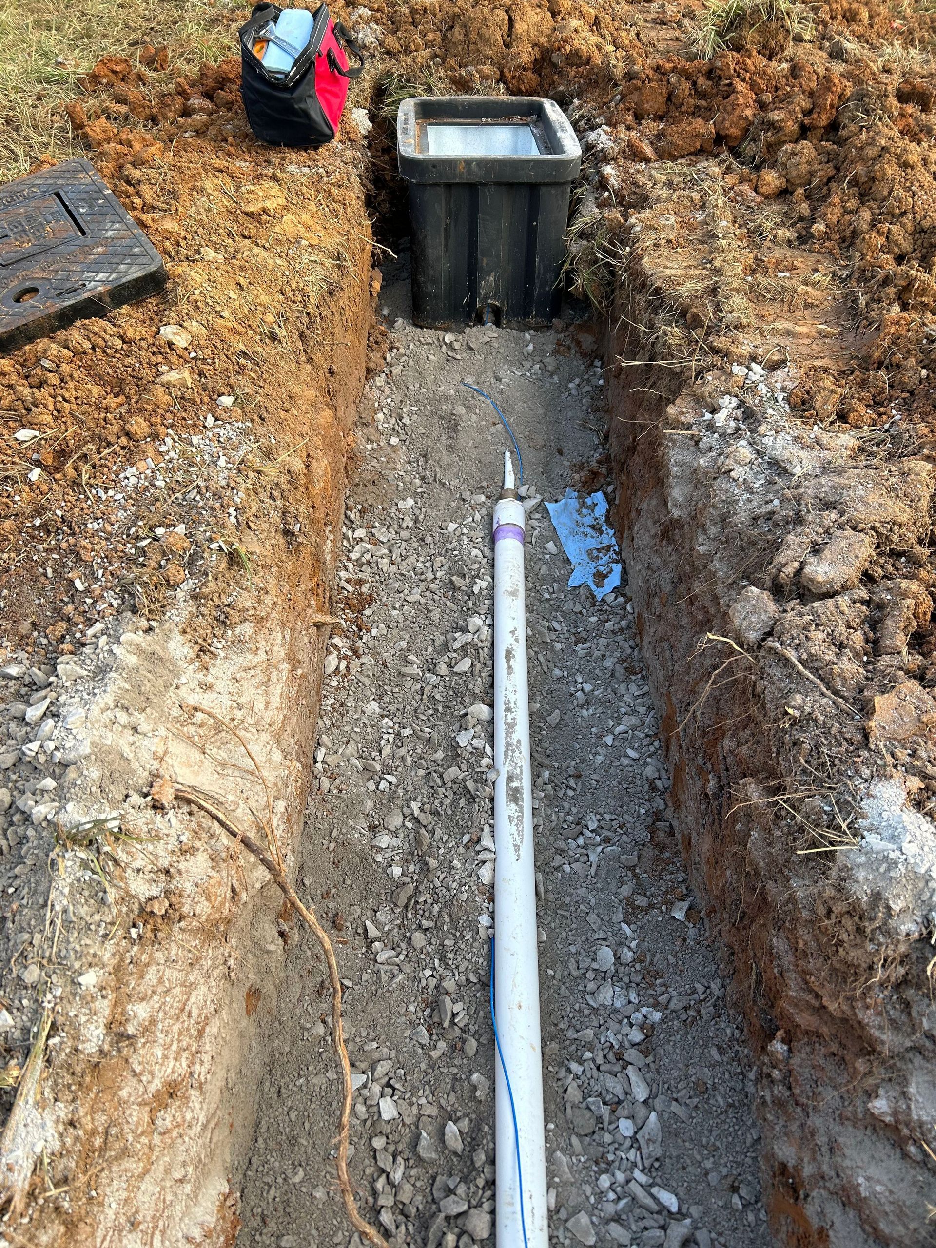 A PVC pipe rests in a gravel-lined trench leading to a rectangular black drainage catch basin in an outdoor setting.
