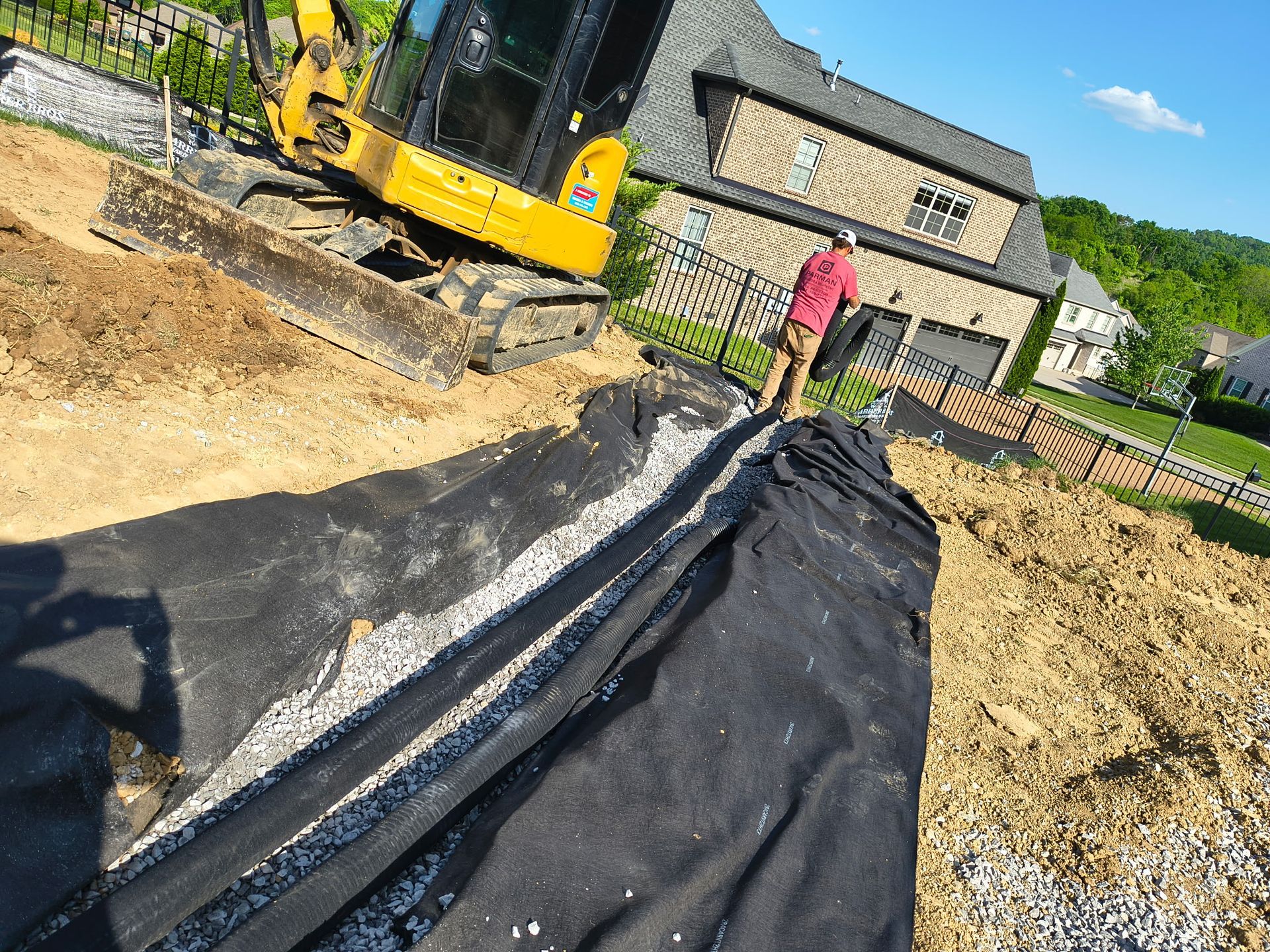 Backyard excavation with black landscape fabric, a worker, and a yellow excavator beside a house