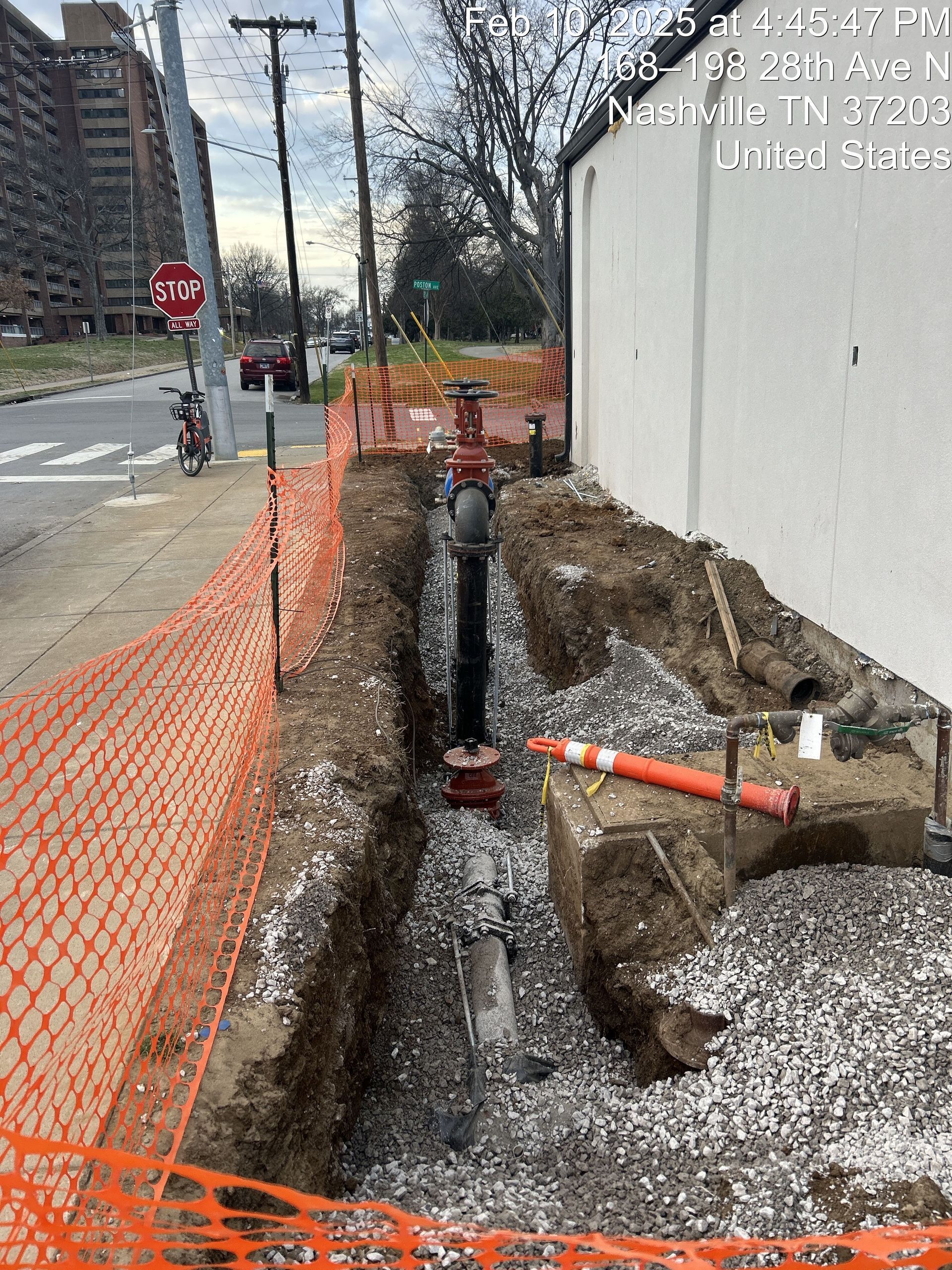 An excavated trench containing utility pipes and gravel runs alongside a white building, cordoned off by orange fencing.