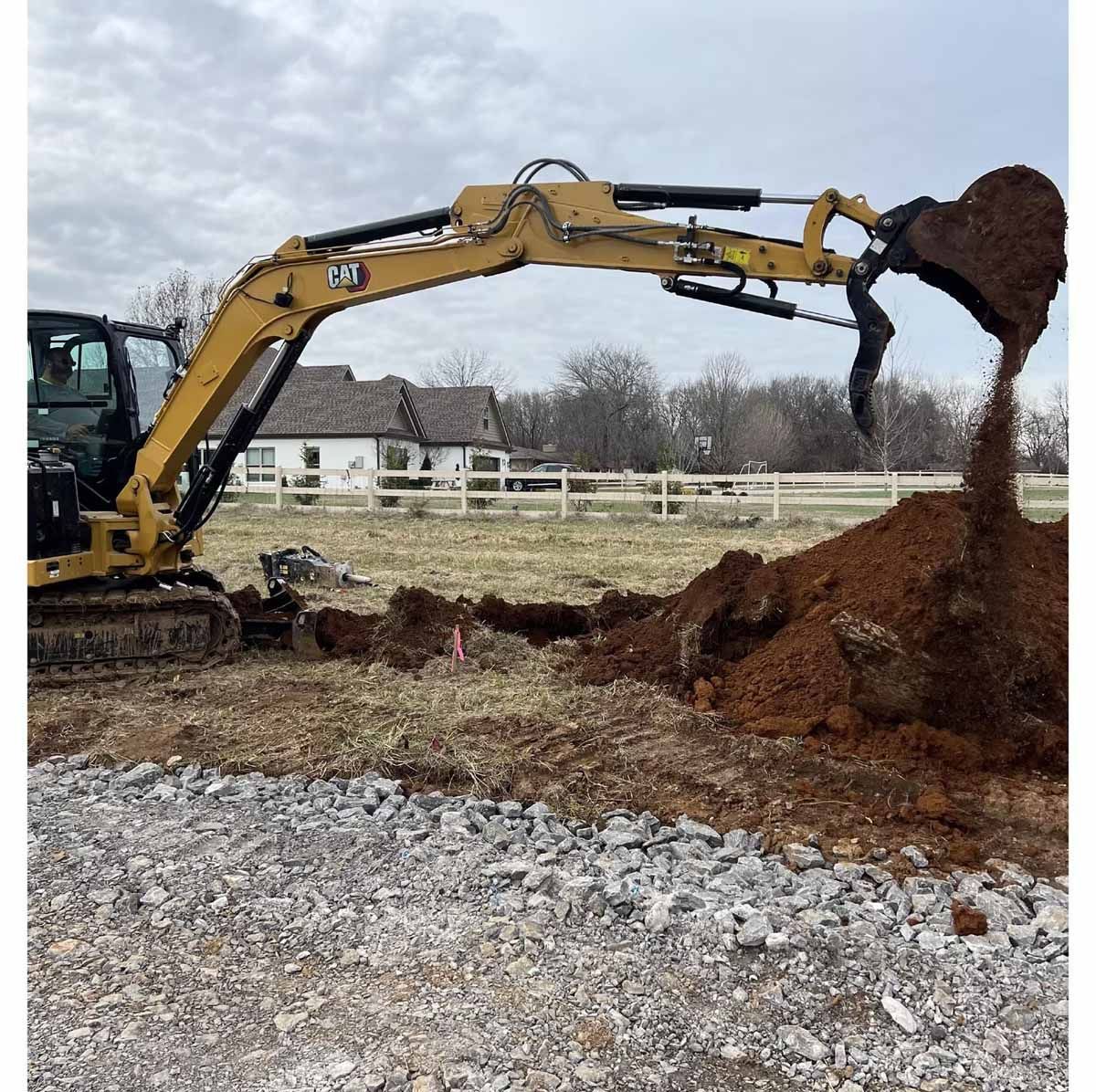 Yellow excavator digging in a field, dumping dirt. White fence and house in the background.