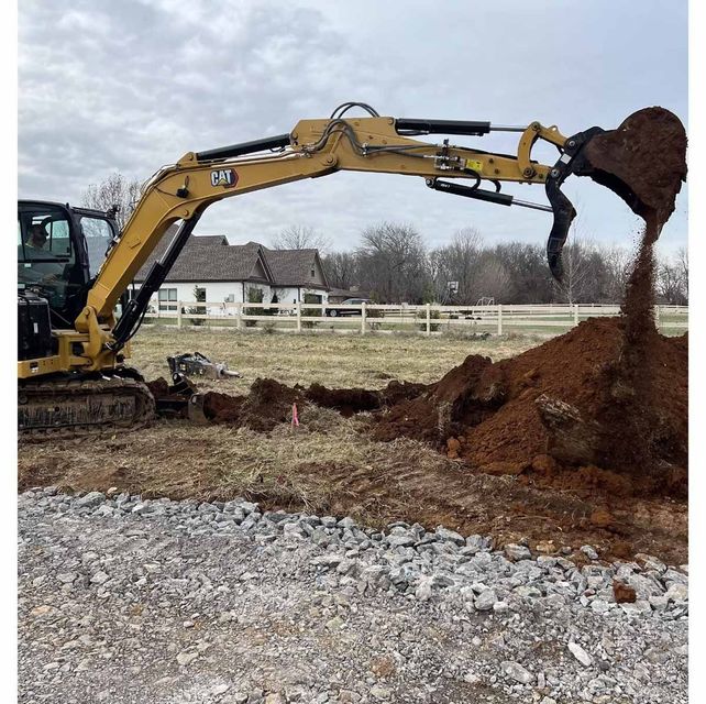 Yellow excavator digging soil in a field near a house and fence.