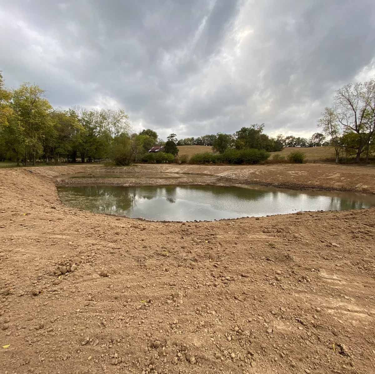A muddy pond surrounded by bare earth under a cloudy sky, trees in the background.