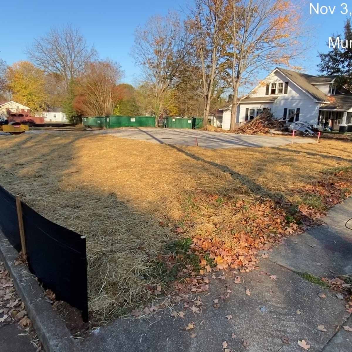Open, grassy lot with a black fence, construction equipment, and a house in the background.