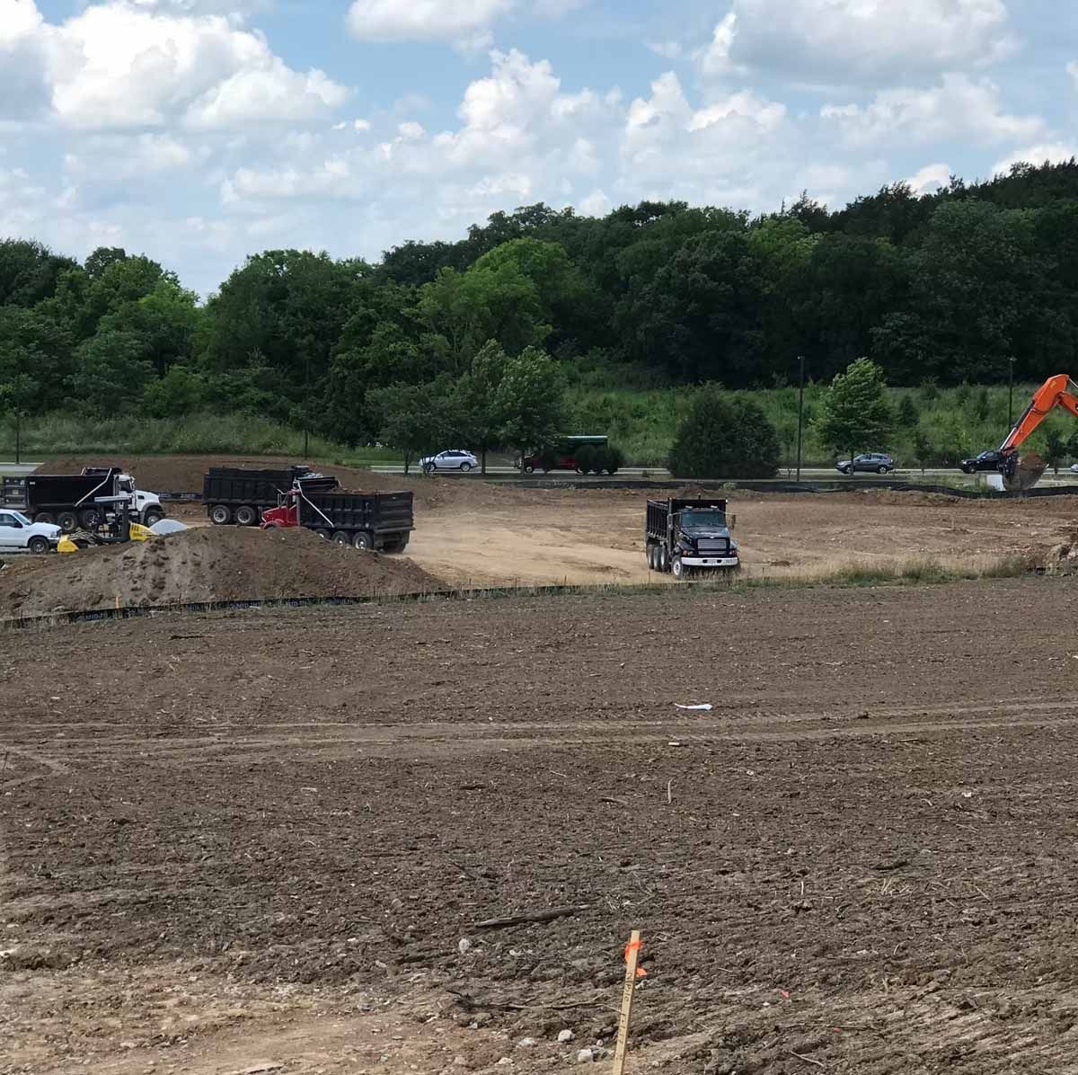 Construction site with dump trucks, excavator, and workers. Dirt field with green trees and blue sky.