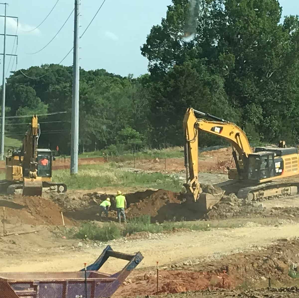 Three yellow excavators on a grassy patch near a small building and trees; some excavation work is underway.