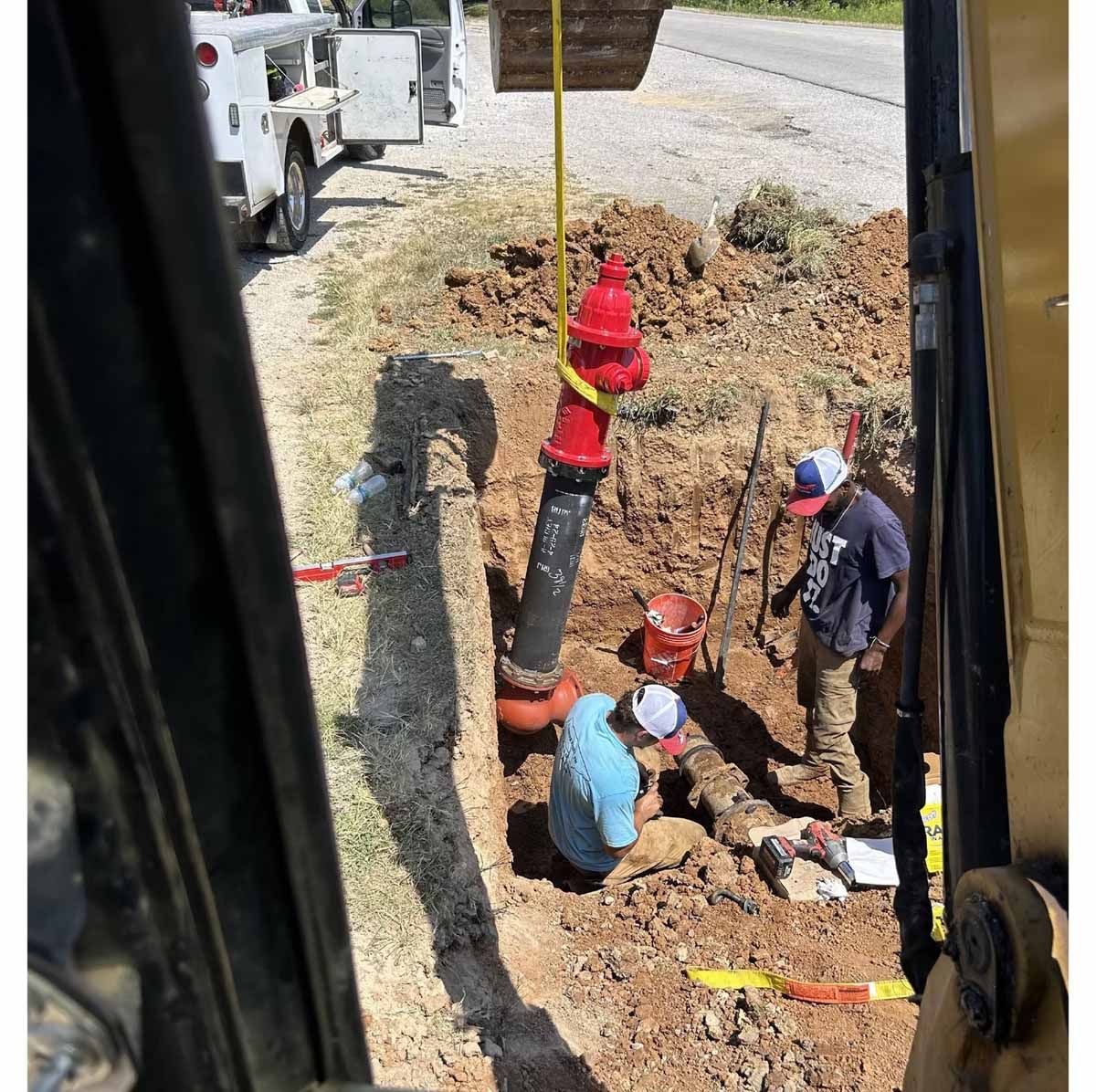 Workers install a red fire hydrant in a dirt pit next to a road.