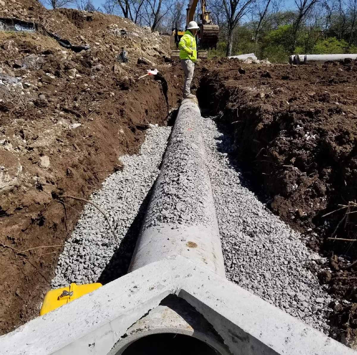 Construction site with concrete pipe and gravel in a trench, worker in the background, sunny day.
