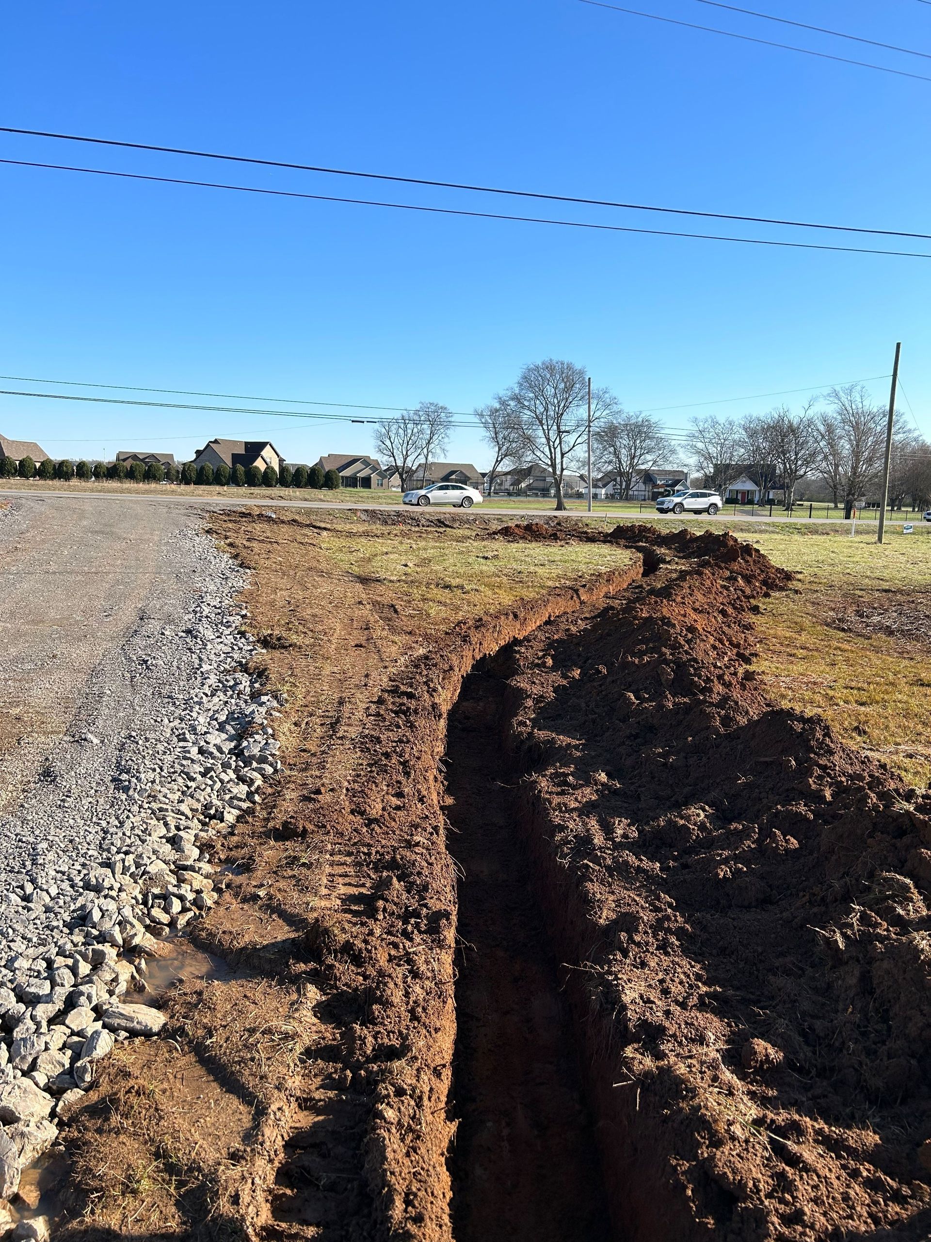 A long, freshly dug dirt trench runs parallel to a gravel road under a clear blue sky.