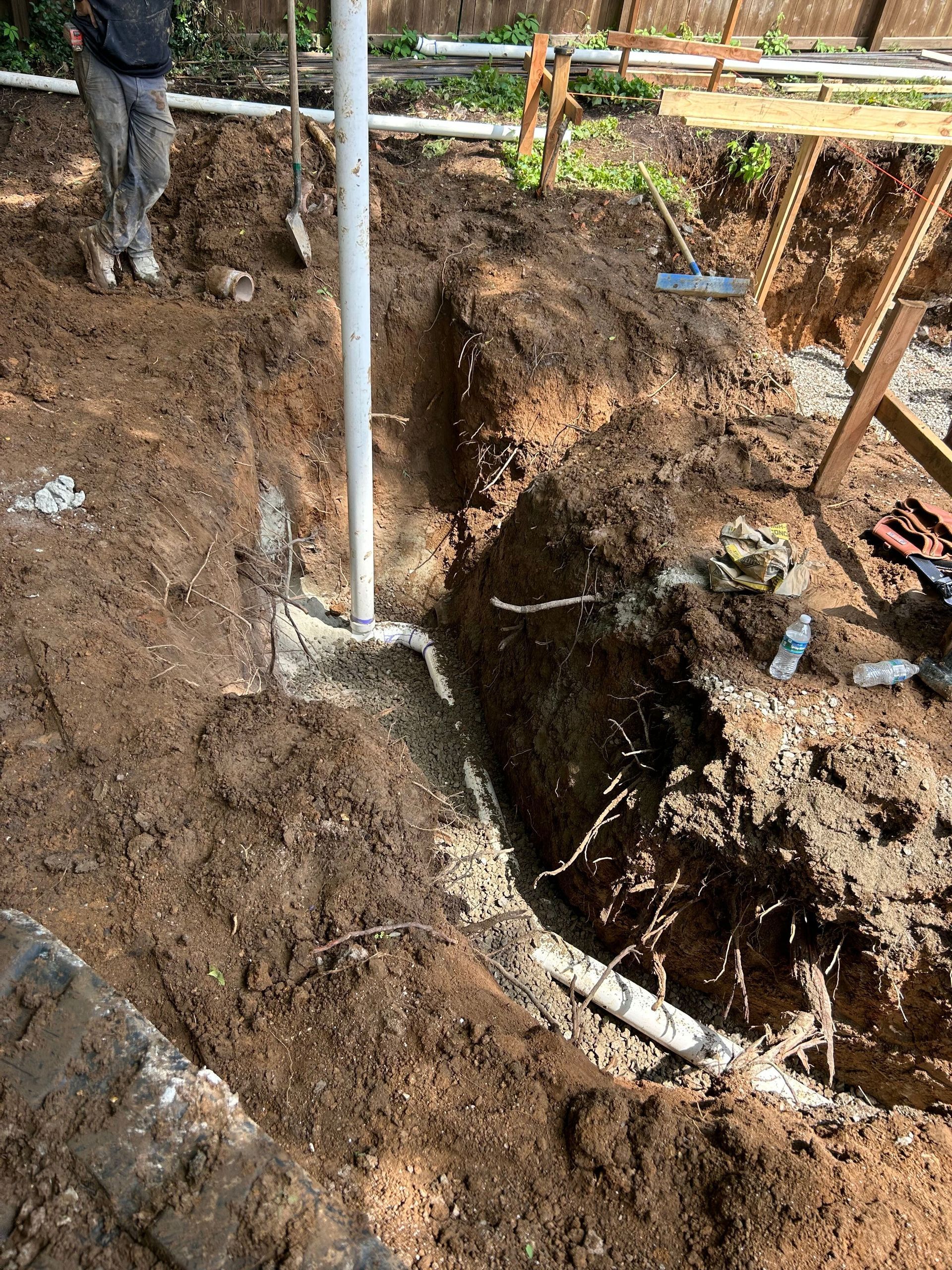 A person stands near a deep trench with exposed white PVC drainage pipes and gravel in a residential backyard.