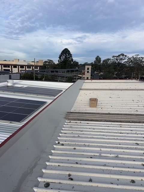 View of a white corrugated metal roof with a solar panel array and a building in the background under a cloudy sky.