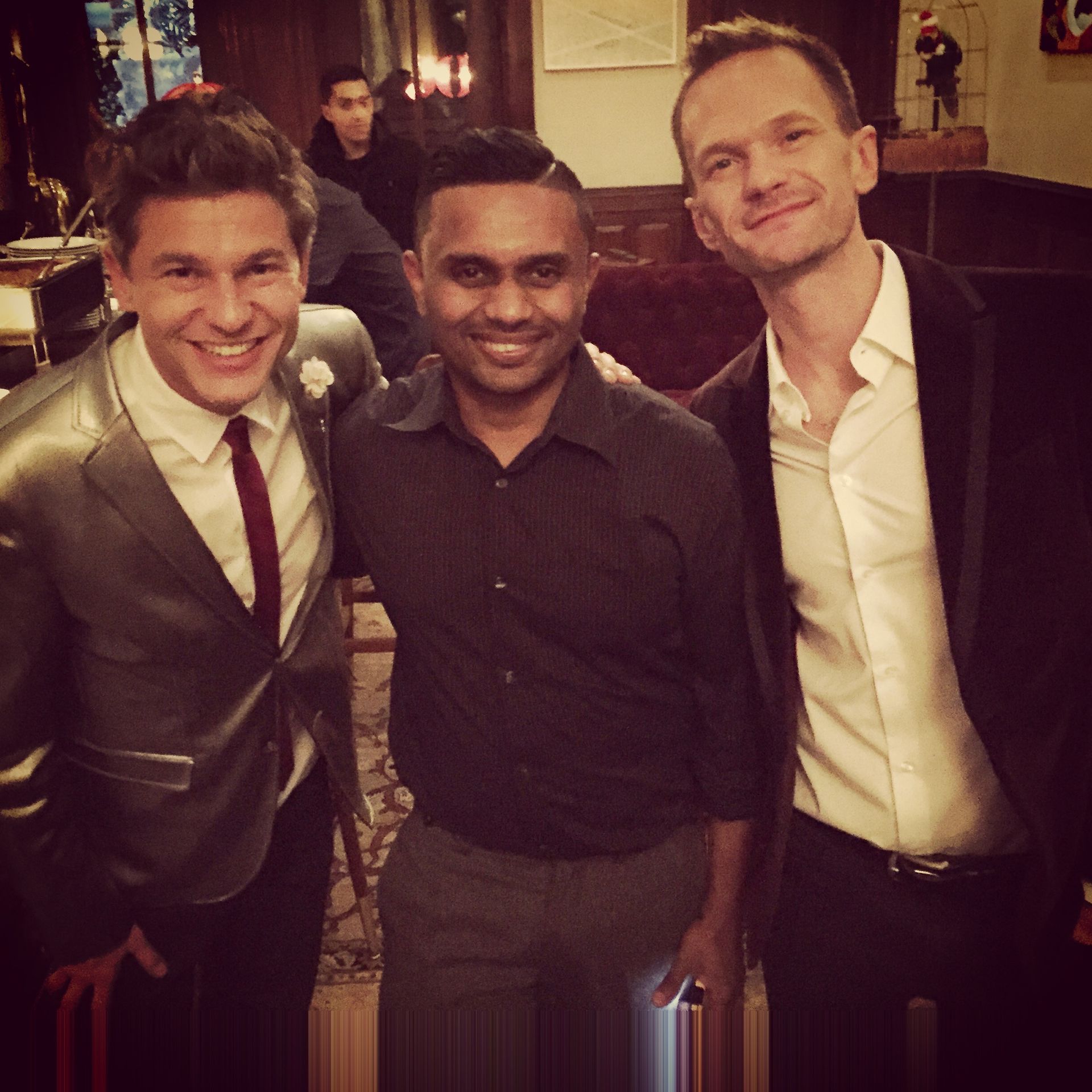 Three men smiling, posing closely together indoors. One in a silver suit, one in a gray shirt, one in a black suit.