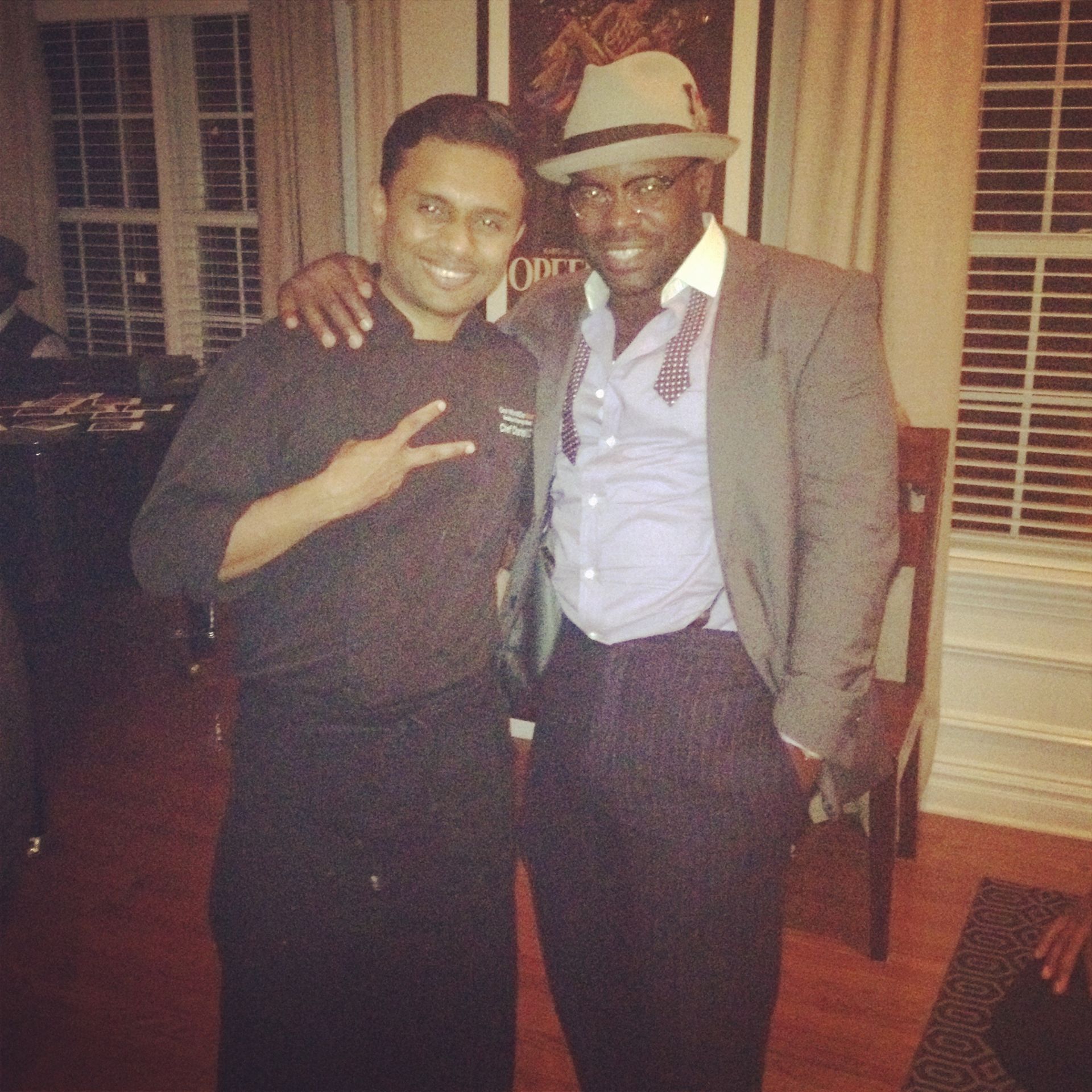 Two men pose. One in chef attire, peace sign. The other, a suit, hat, arm around the chef, indoors.