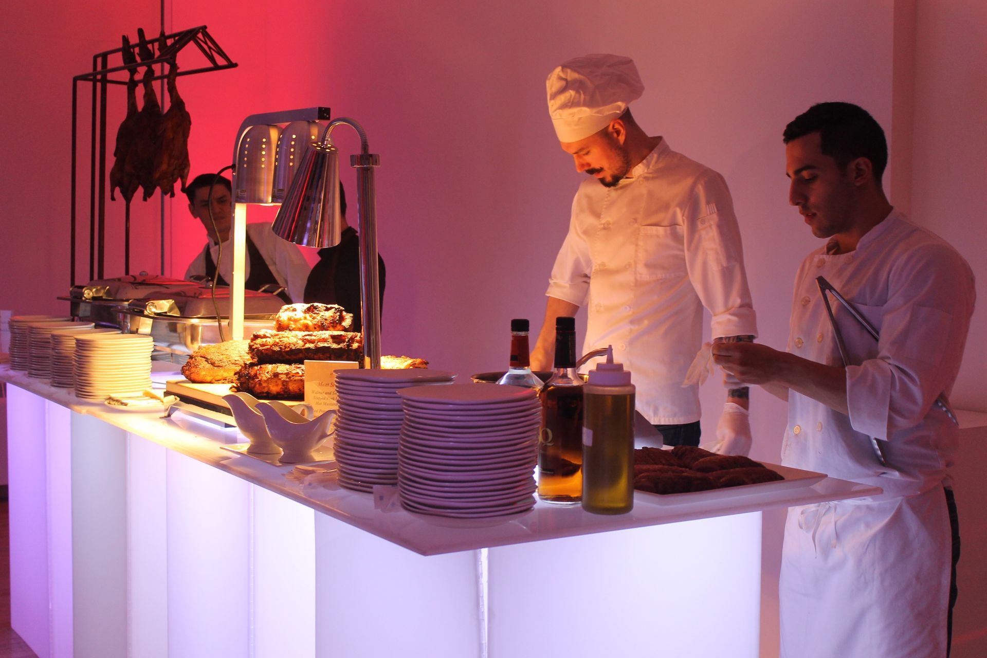 Chefs preparing food at a buffet. Brightly lit buffet table with dishes, sauces, and hanging meats.