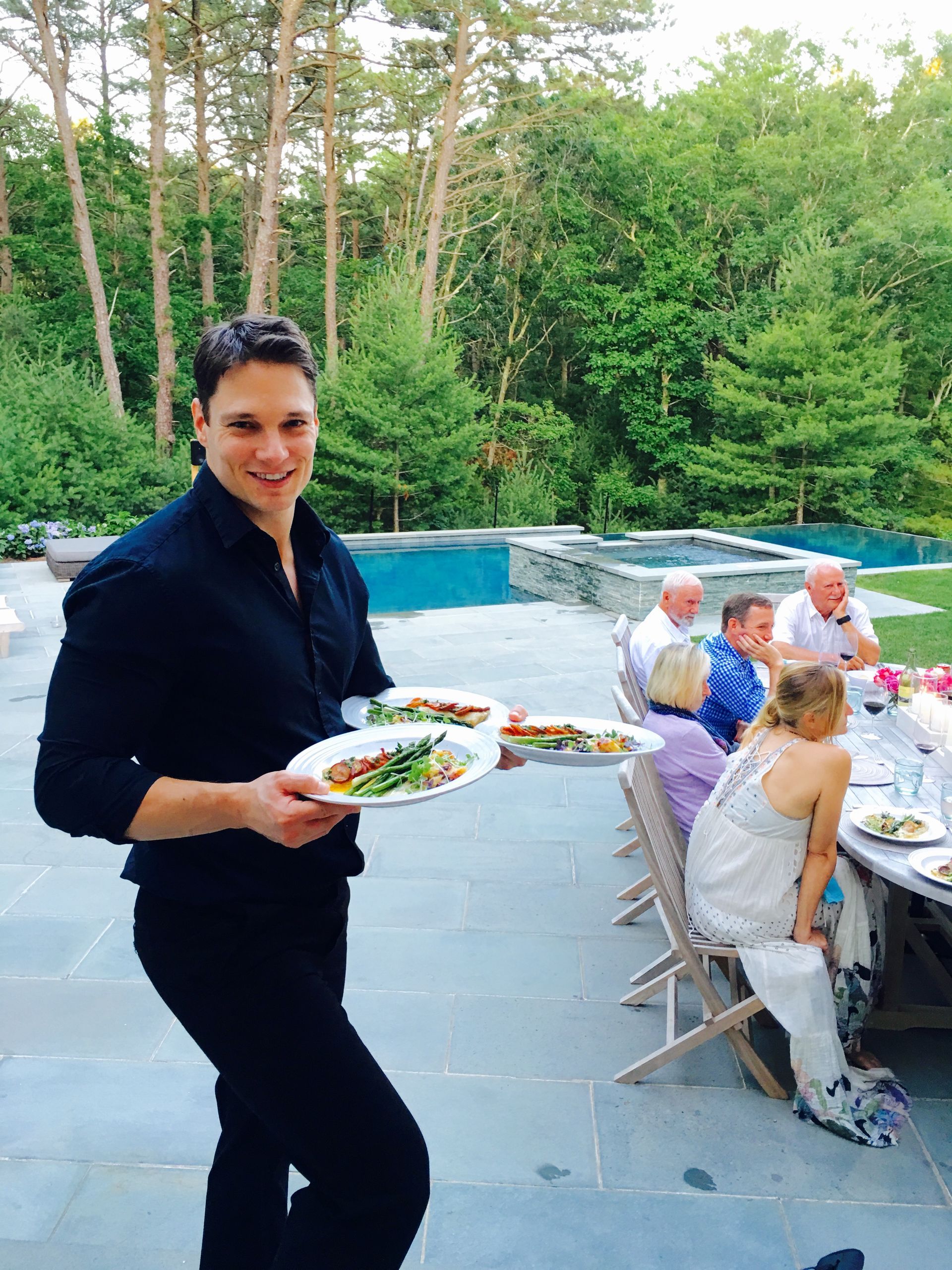 Server holding plates of food outside; people seated at a table, pool and trees in background.