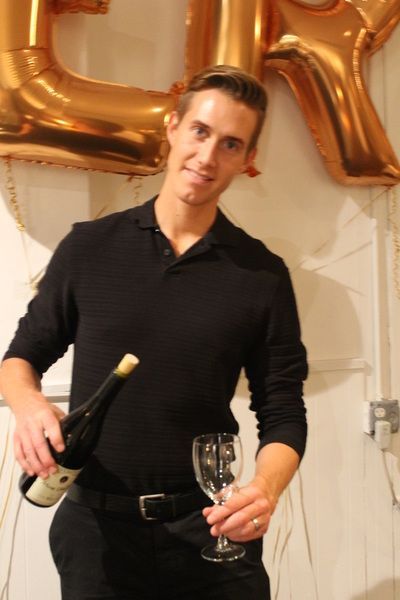 Man pouring wine into a glass; gold balloons in background, black outfit.