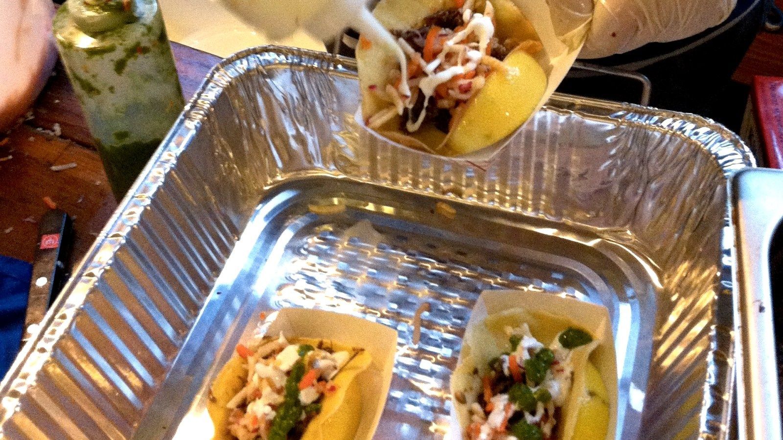 Tacos being placed into a foil pan. The tacos have fillings and garnishes; cilantro, and white sauce.