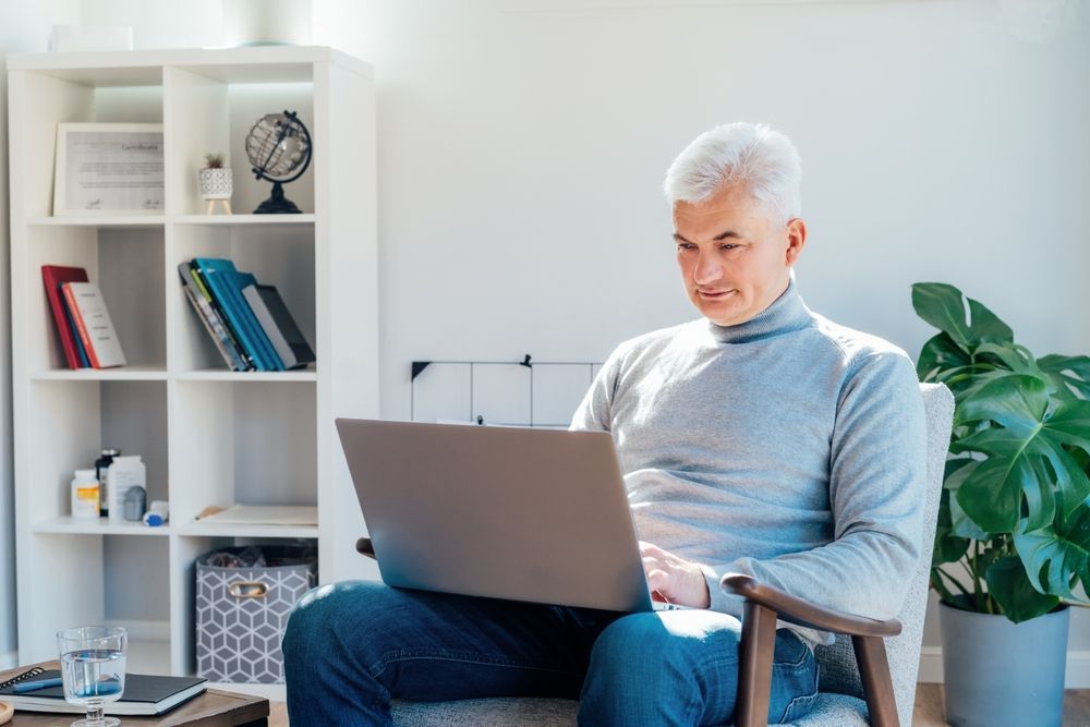 Man with gray hair, using a laptop, sitting in a chair, indoors. A bookshelf and plants are nearby.