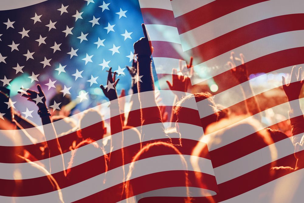 American flag overlaying a crowd with raised hands, possibly at a concert or rally.