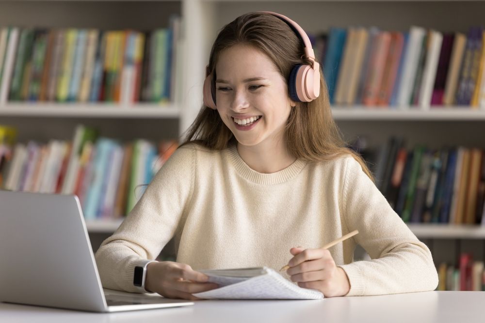 Woman wearing headphones, smiling at a laptop, taking notes in a library setting.